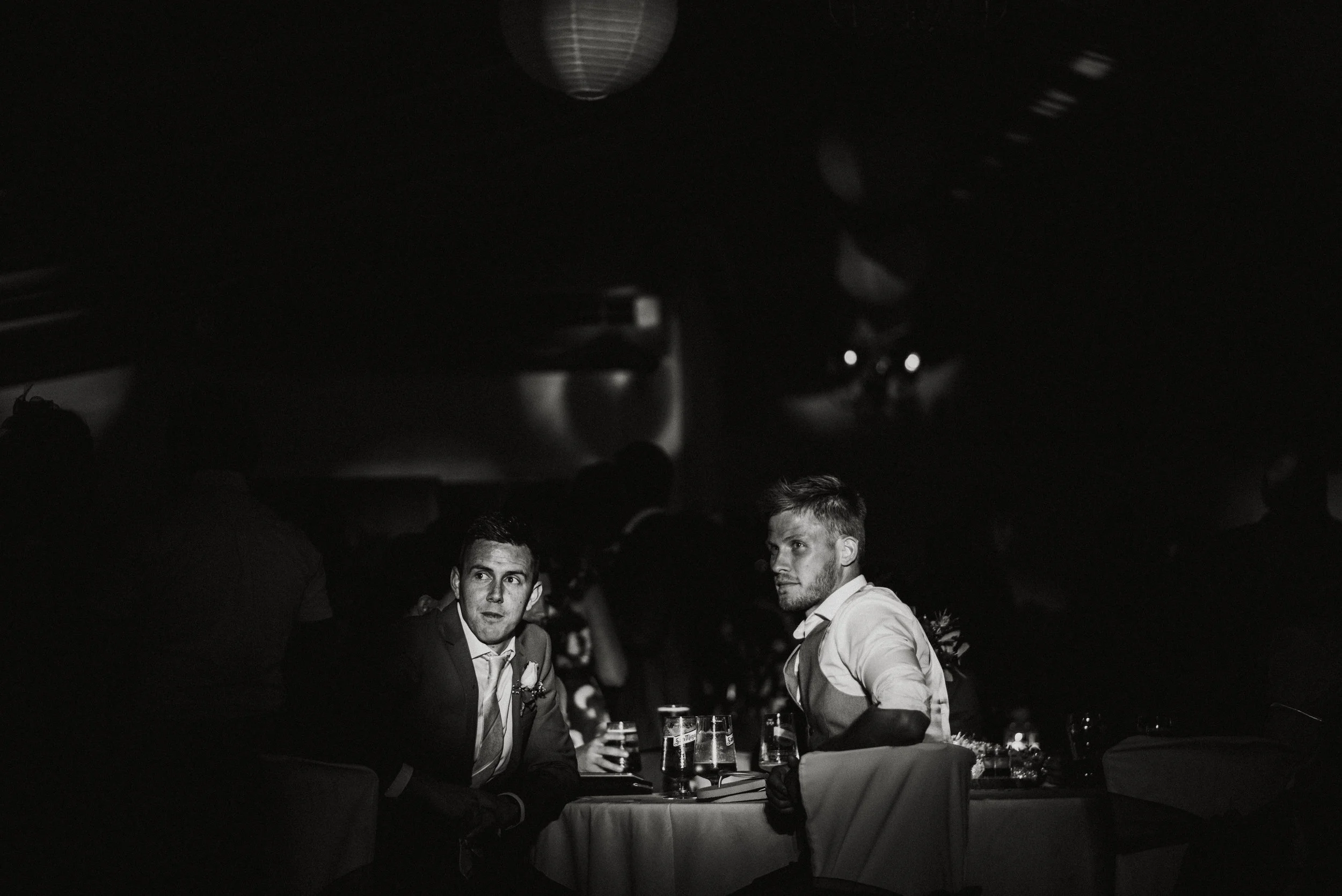 Two men sitting at a table in a dark, elegant setting, likely at a formal event, with drinks in front of them.