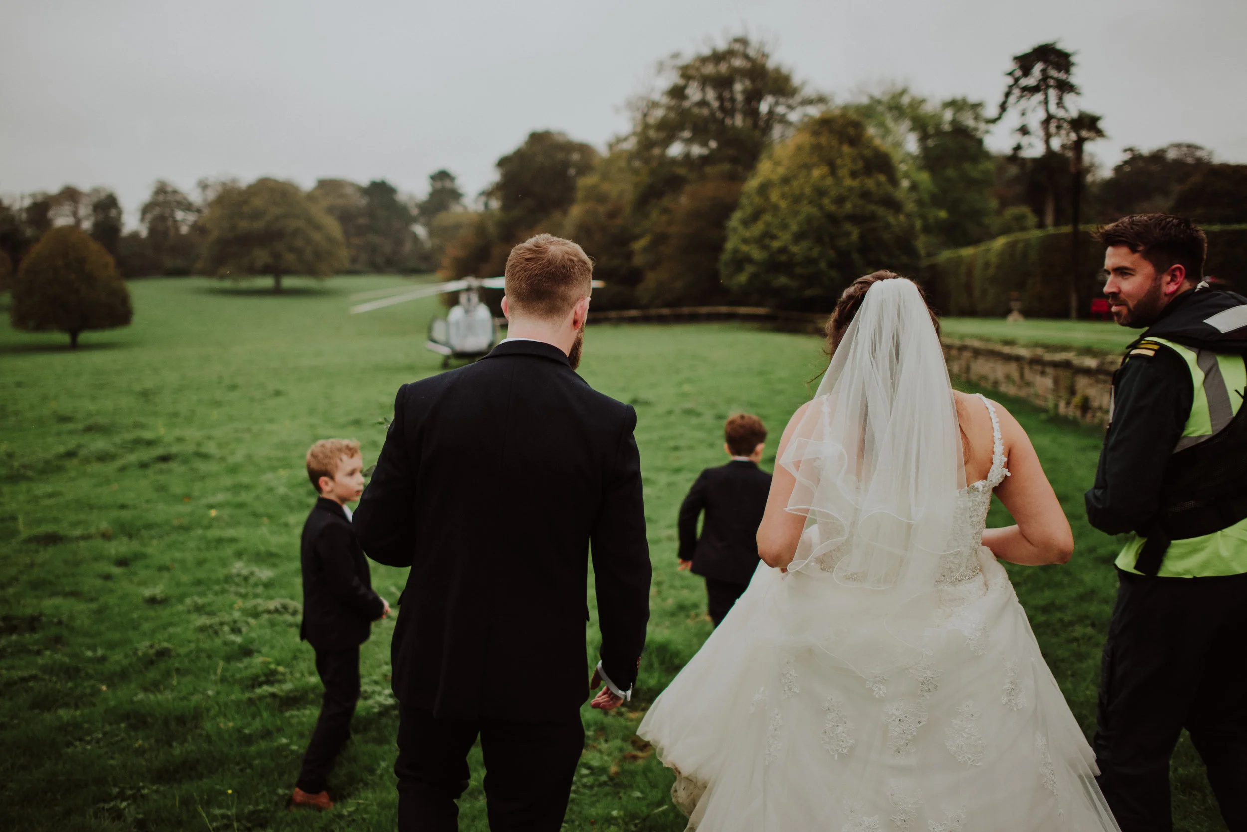 A bride in a white wedding dress and veil holding hands with a groom in a black suit, walking on a grassy field towards a helicopter in the background, with a man in a high-visibility vest and three young boys also in suits.