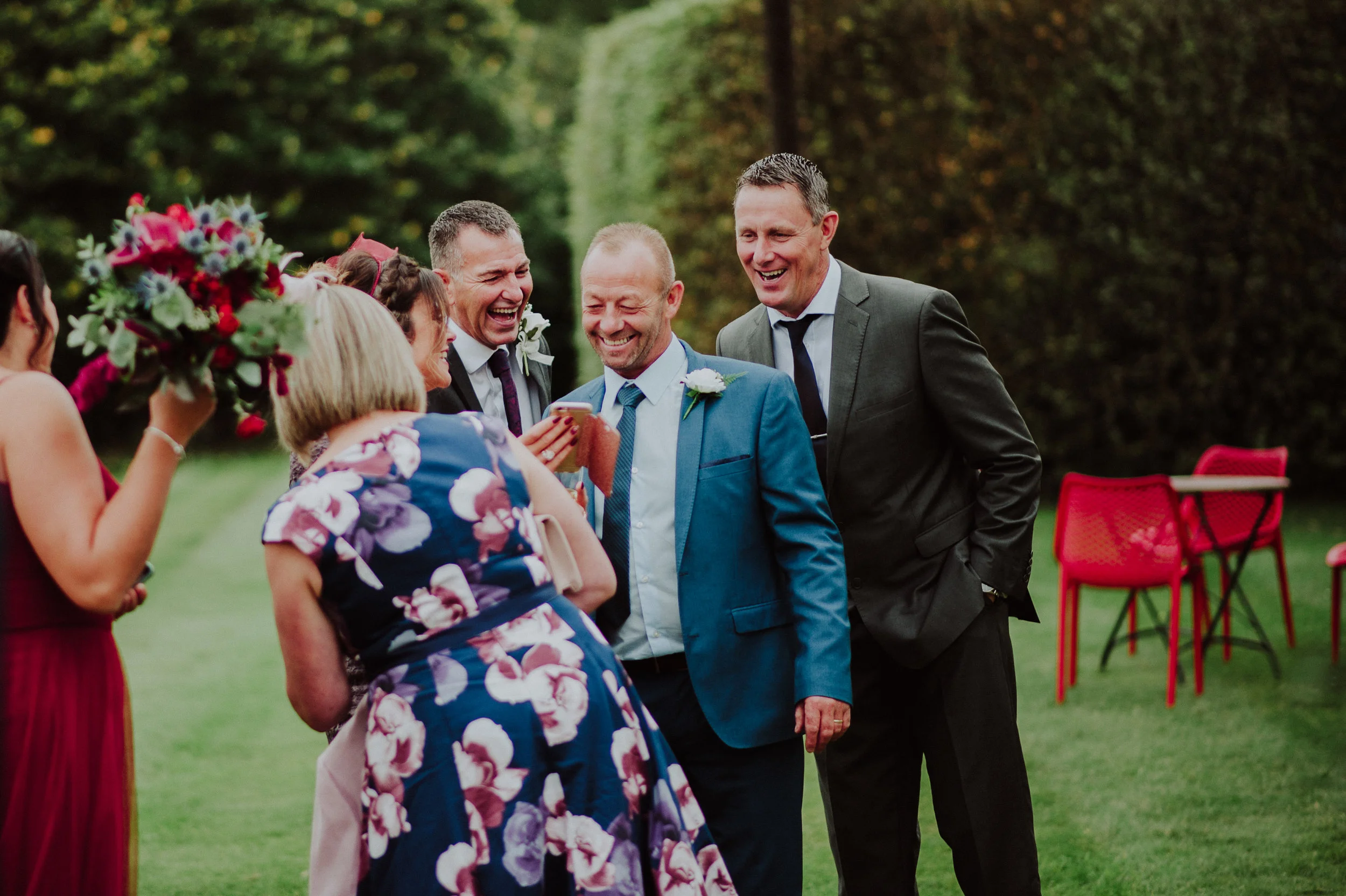 A group of people dressed in formal attire, including men in suits and women in dresses, are gathered outdoors in a joyful moment, smiling and interacting with each other during a celebration, with trees and red chairs in the background.