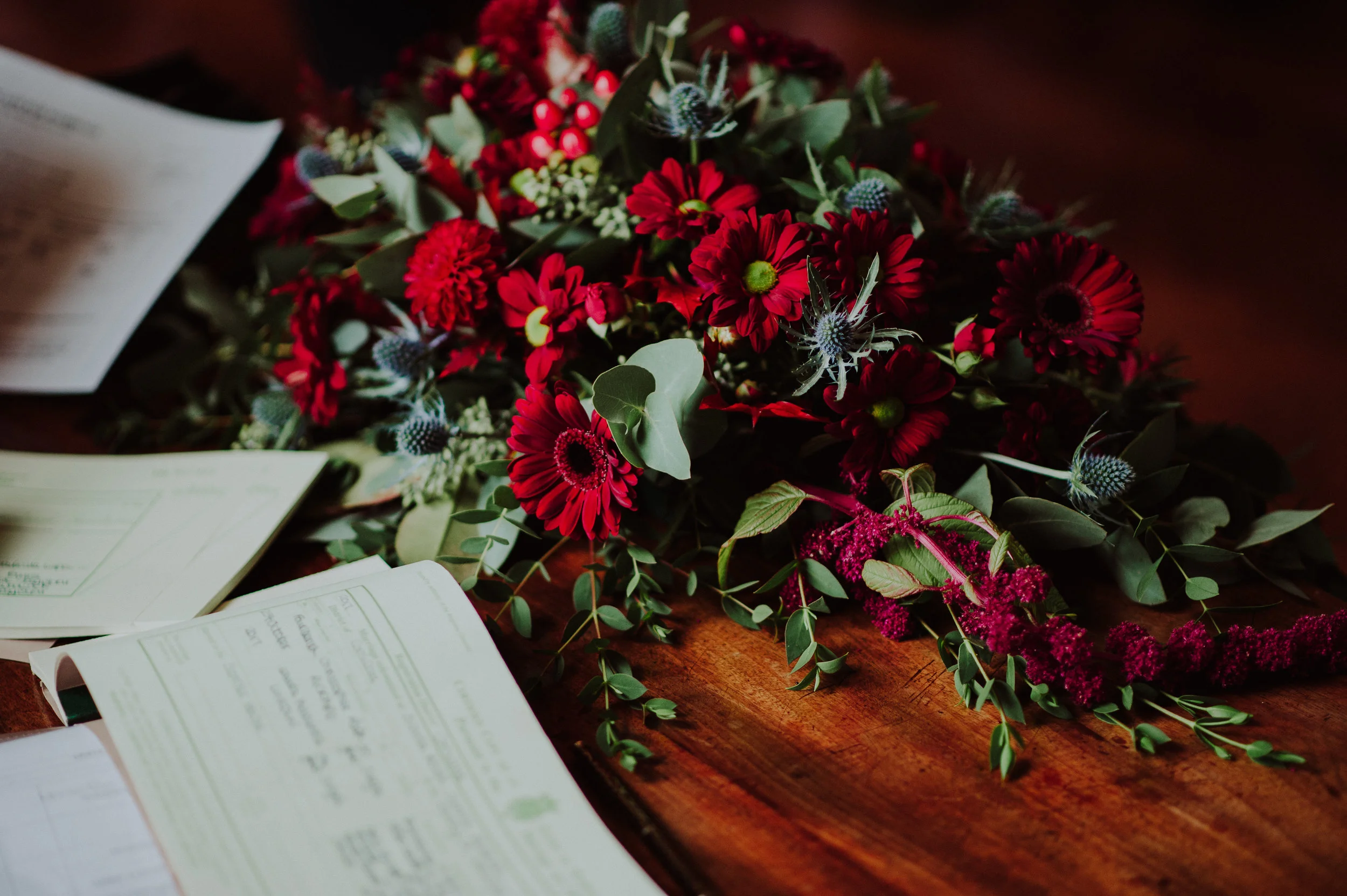 A colorful floral arrangement with red, purple, and green flowers and foliage, surrounded by papers on a wooden surface.