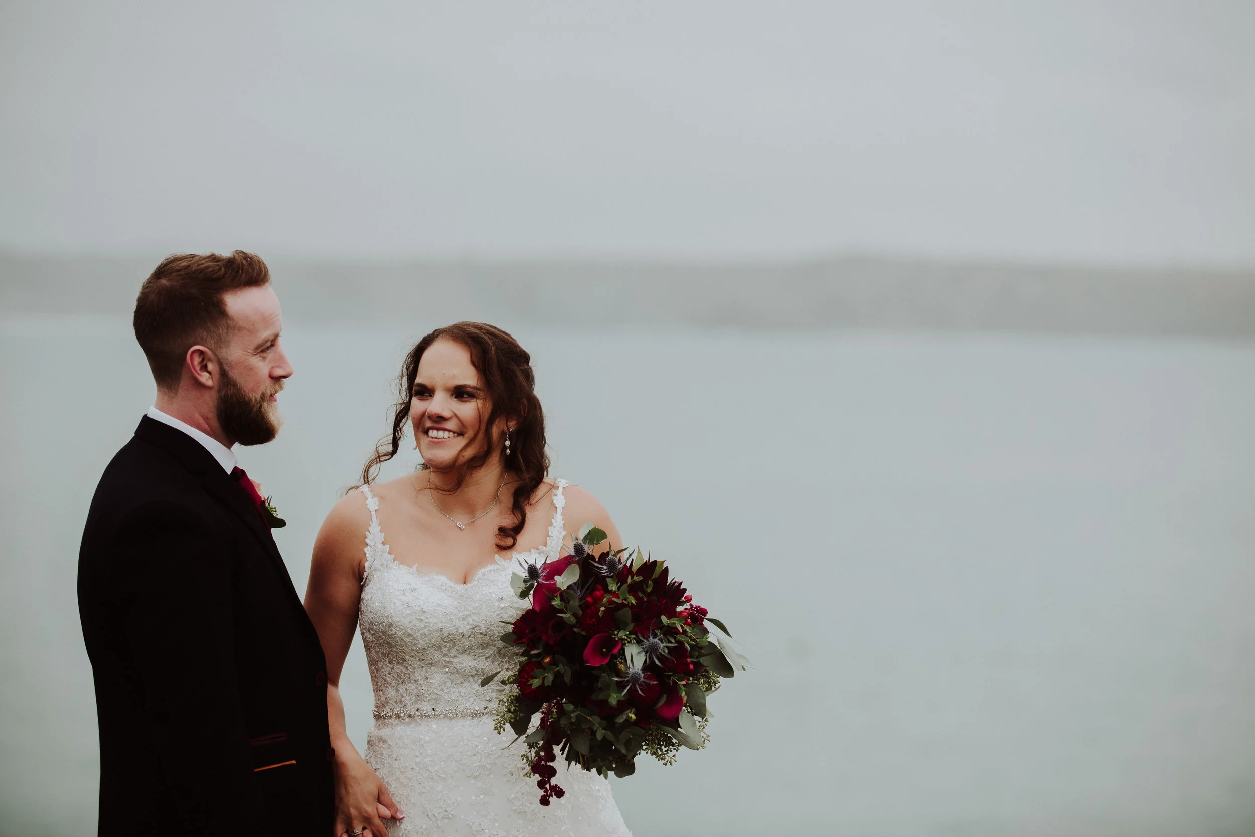 A bride and groom holding hands and smiling at each other near a body of water, with the bride holding a bouquet of dark red and green flowers, on an overcast day.