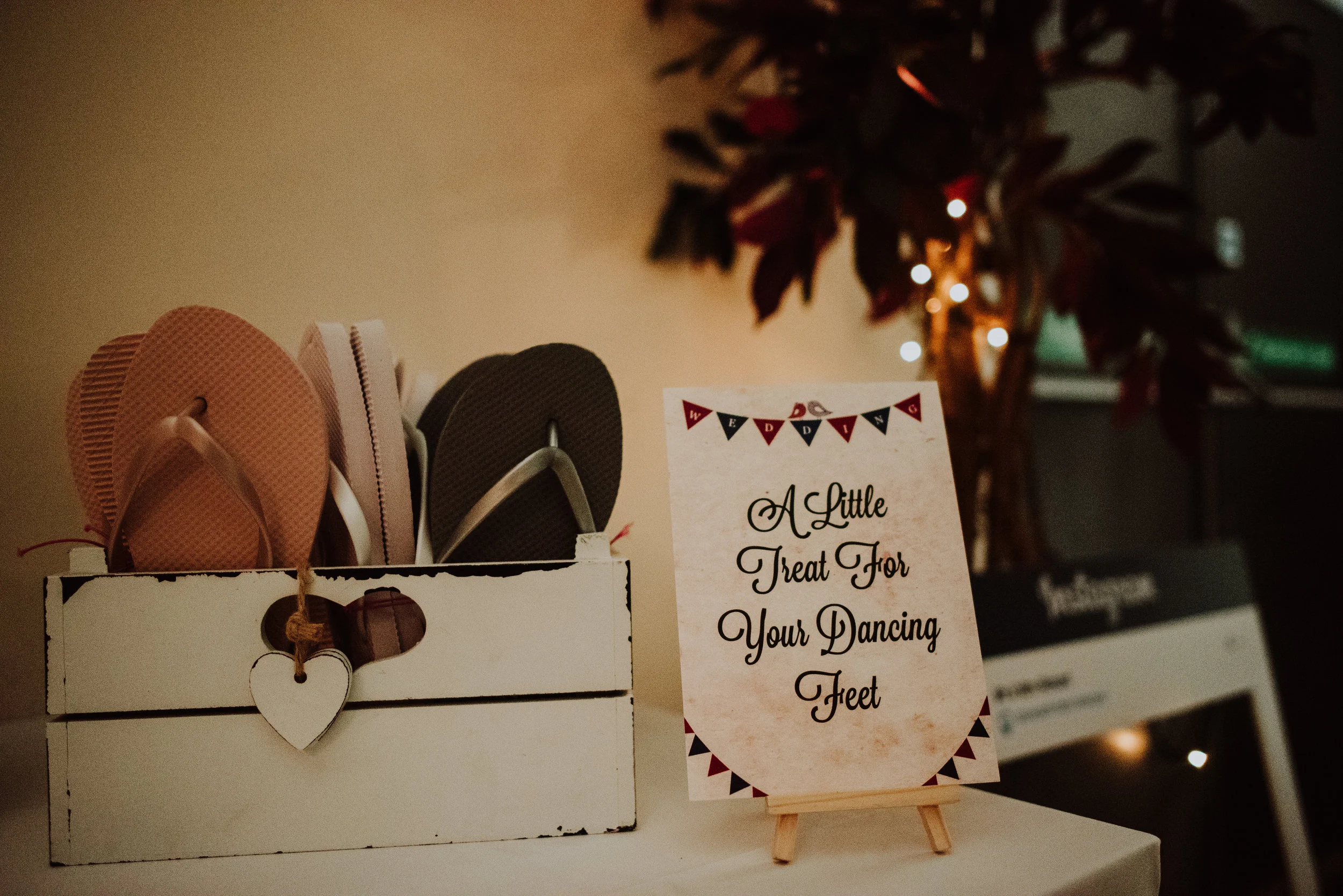 A collection of pink and black flip-flops in a white wooden box, with a sign that reads "A Little Treat For Your Dancing Feet" next to it on a small easel, and blurred decorative lights and tree in the background.