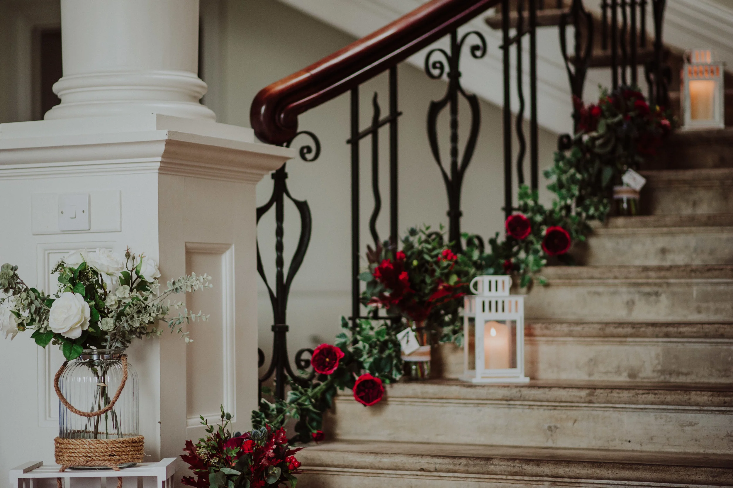 Decorated staircase with red and white flowers, lanterns, and greenery, featuring ornate black railing and white wood trim.