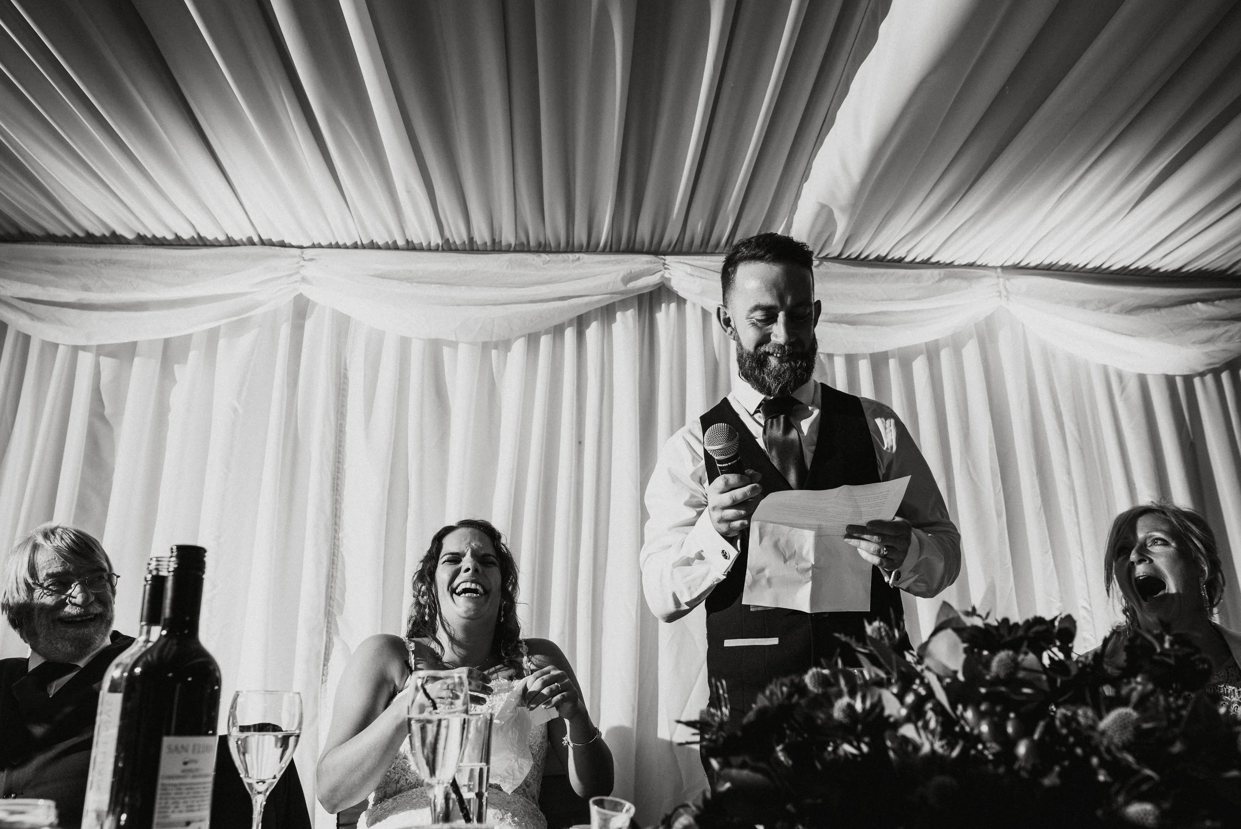 Man in vest and tie giving a speech at a wedding reception, holding a microphone and notes, surrounded by laughing guests.