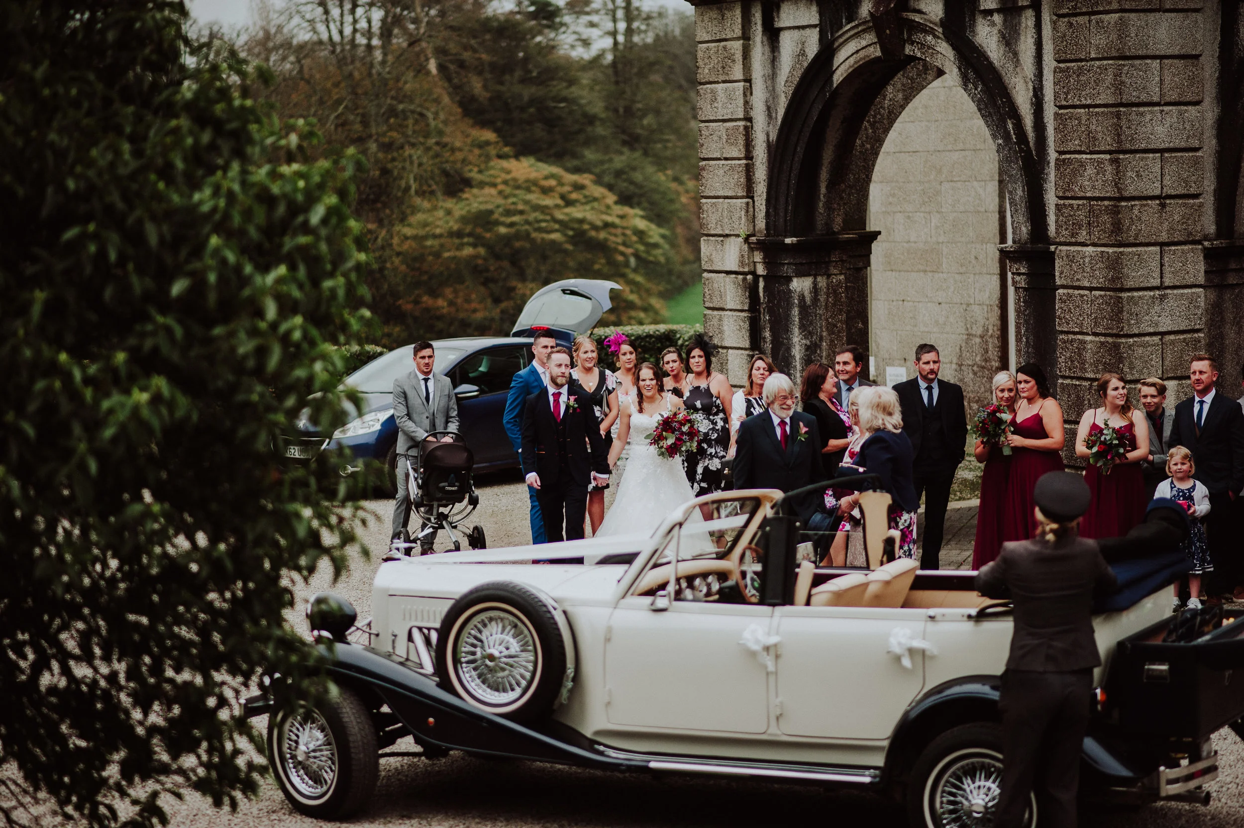 A wedding celebration outdoors with a vintage white car carrying a bride in a white dress holding a bouquet, surrounded by guests dressed in formal attire, near a historic stone building.