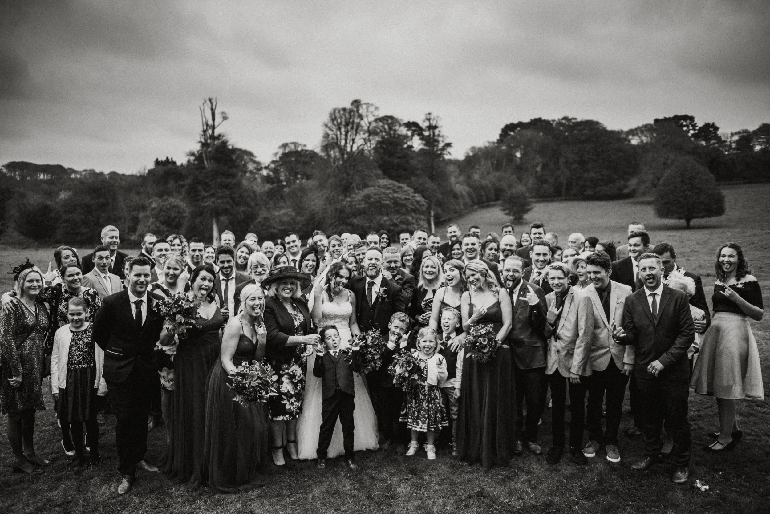 Black and white photo of a large group of people, including children, adults, and some holding bouquets, posing outdoors on a grassy field with trees and hills in the background during a wedding celebration.