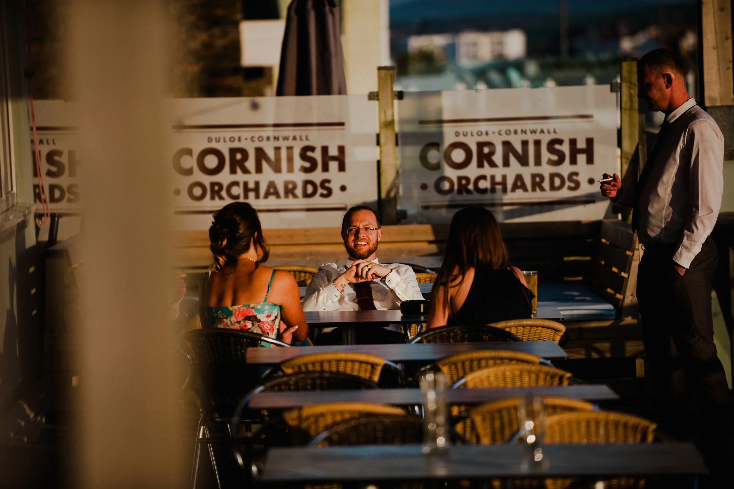 People dining outdoors at a restaurant with a sign that reads 'Cornish Orchards' in the background.