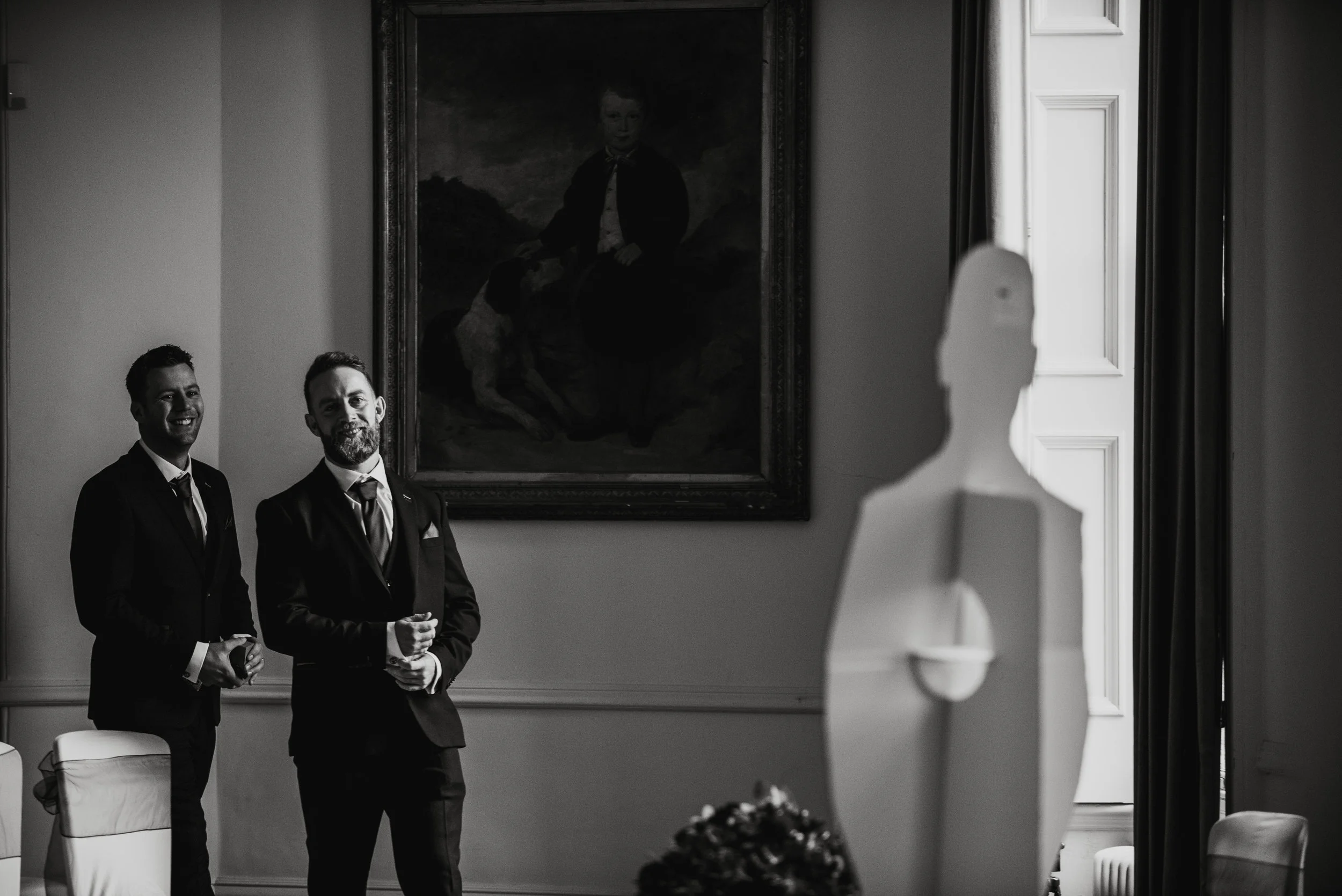 Black and white photo of two men dressed in tuxedos, standing in a room with a large portrait on the wall behind them. A white sculpture in the foreground partially obscures the scene.
