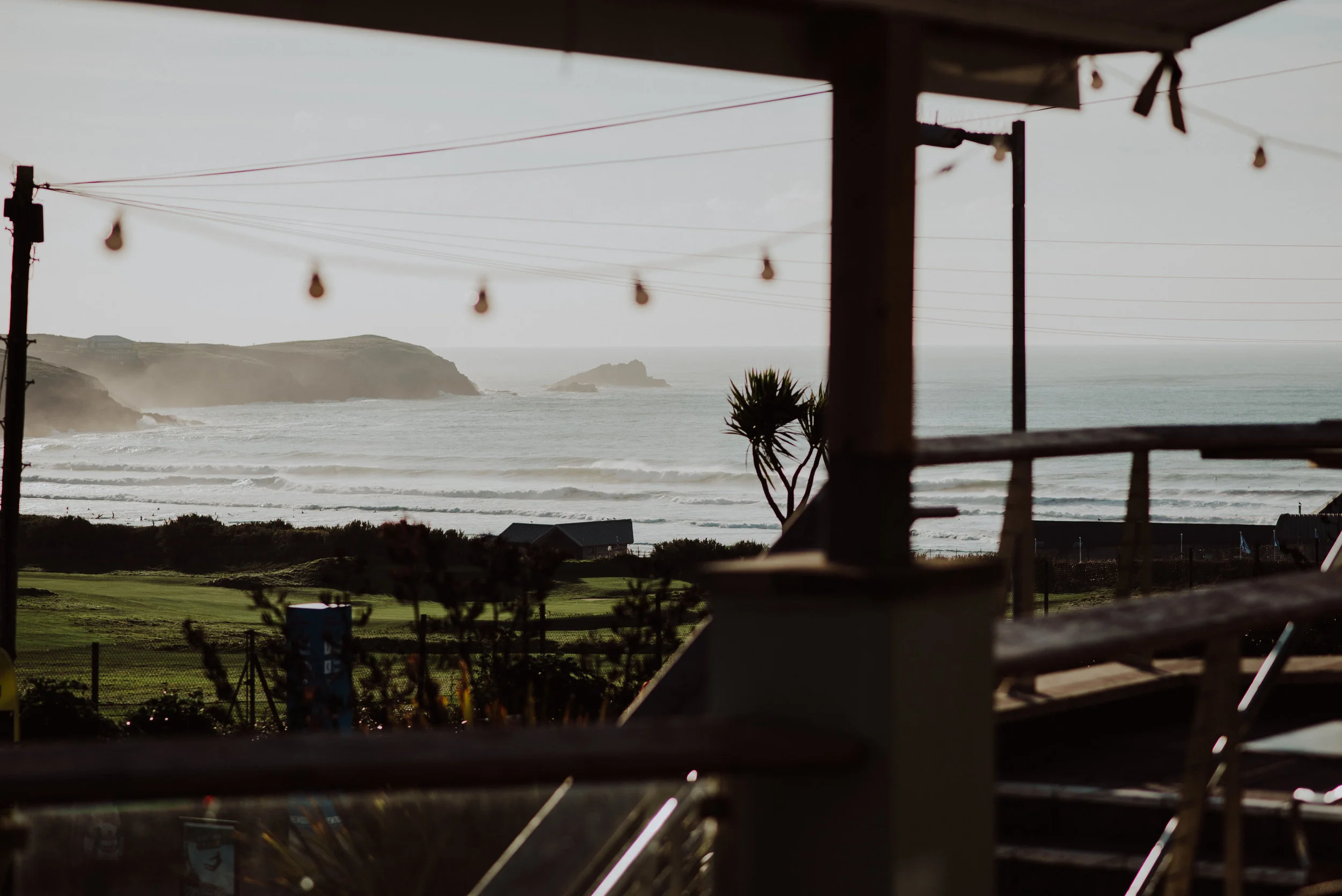 View from a porch or balcony overlooking a beach with waves crashing, distant islands, and hills in the background. Part of the porch's structure and a palm tree are visible in the foreground, along with outdoor string lights hanging above.