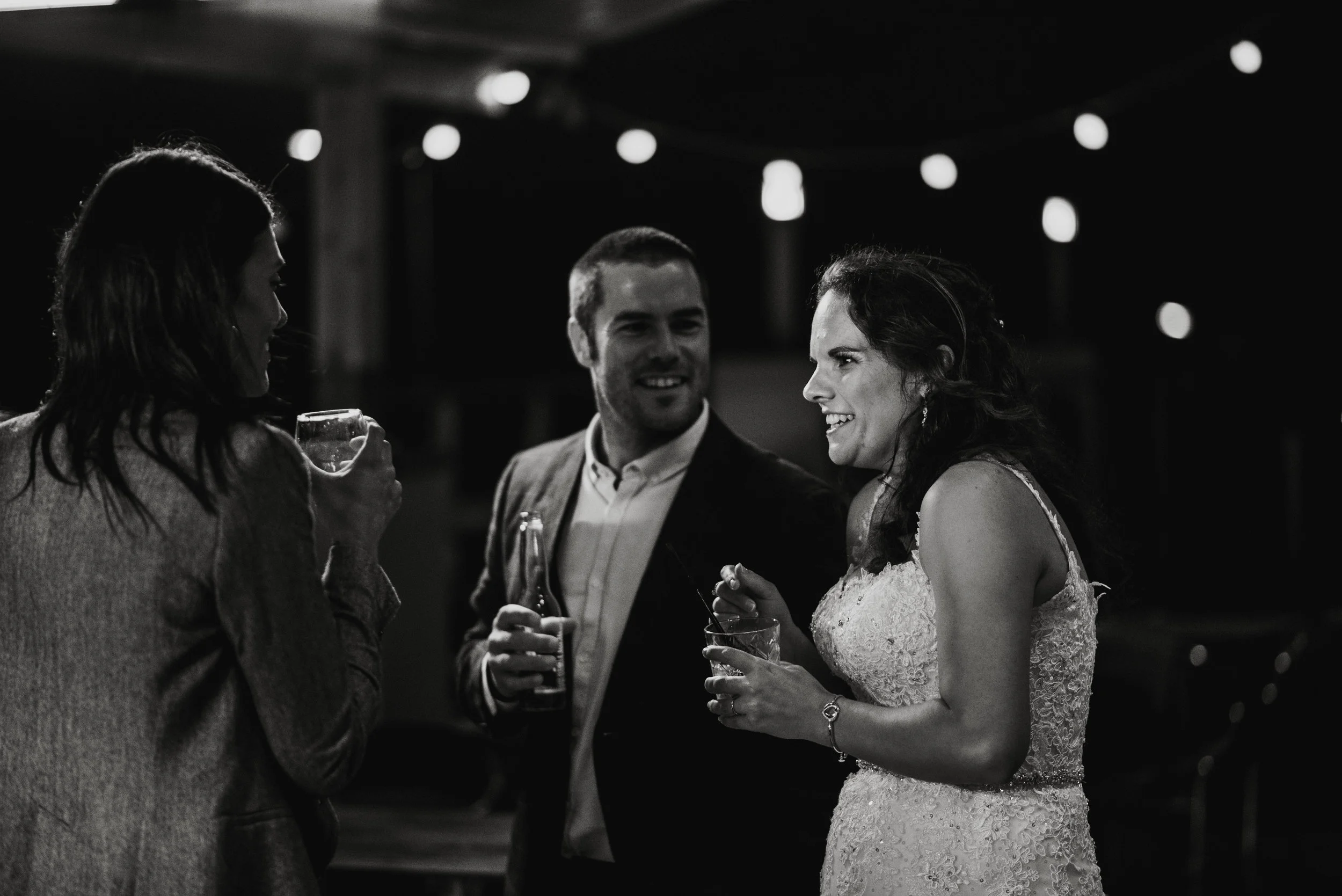 Three people having a conversation at a nighttime event, with string lights in the background. One woman is dressed in a wedding gown, and the other two are dressed in semi-formal clothing, holding drinks.