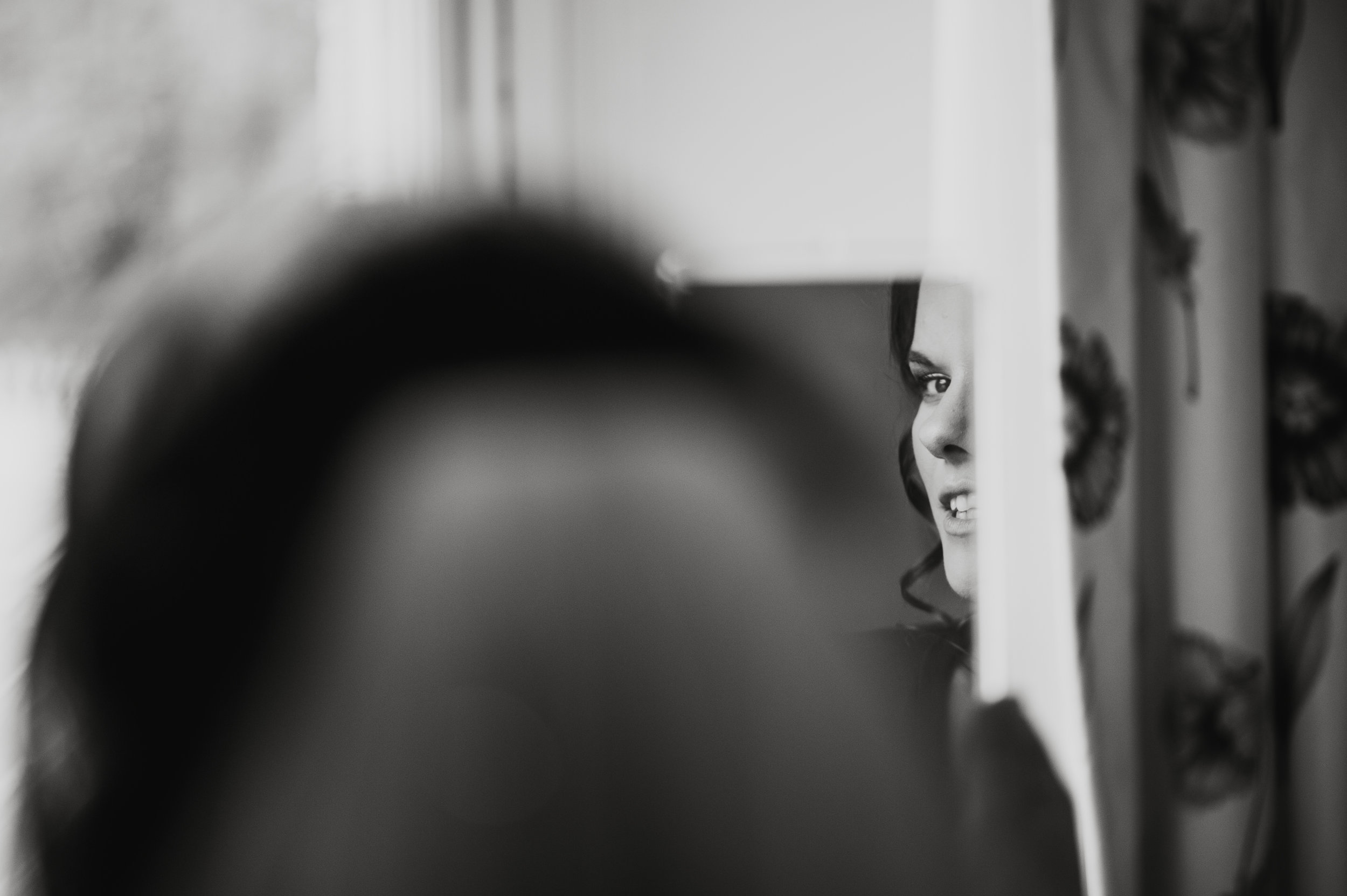 A black and white photo shows a woman with curly hair smiling, seen through a small opening or gap in a piece of furniture or structure.
