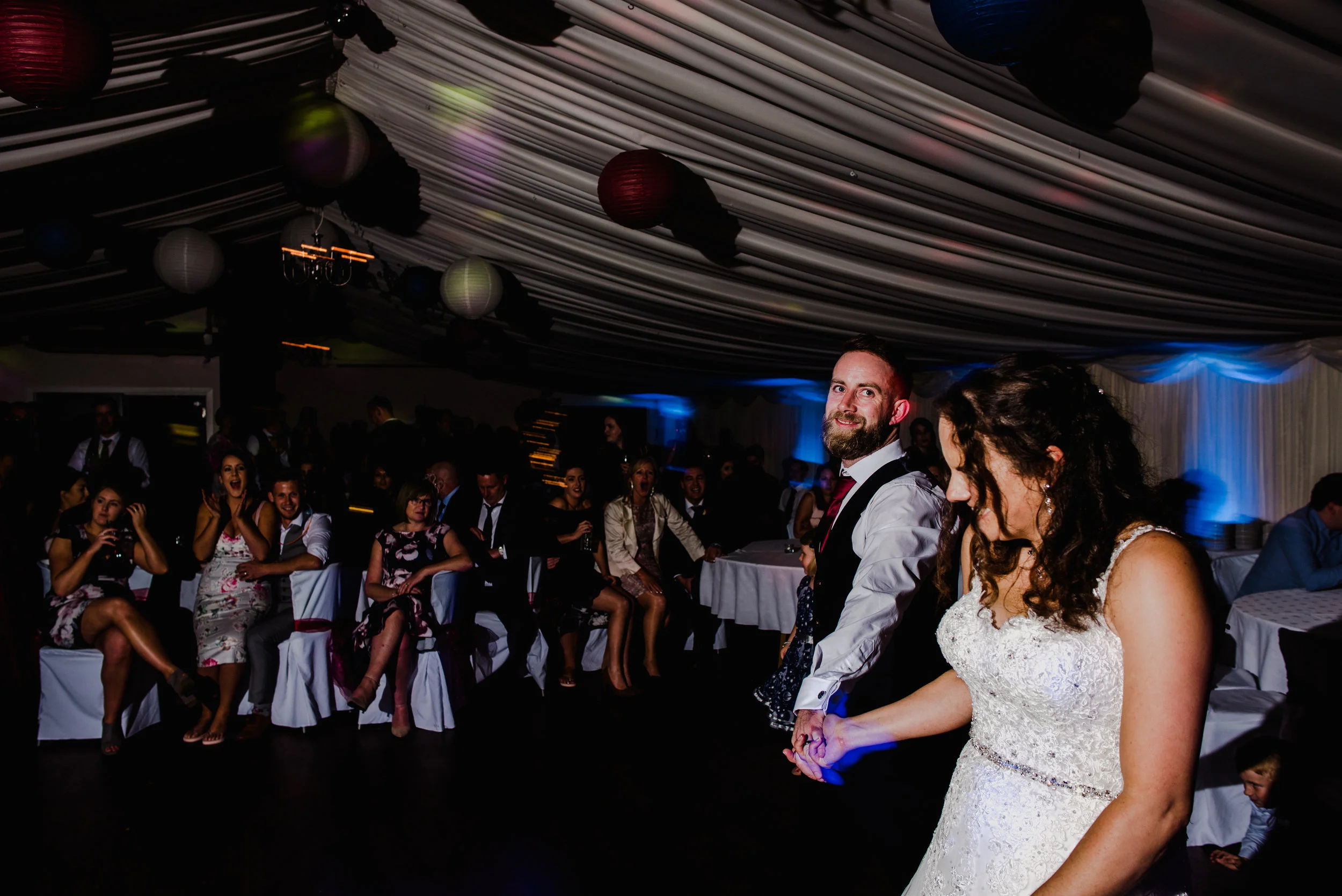 A bride and groom holding hands during their wedding dance, with guests seated and watching in the background under a decorated ceiling with hanging lanterns.