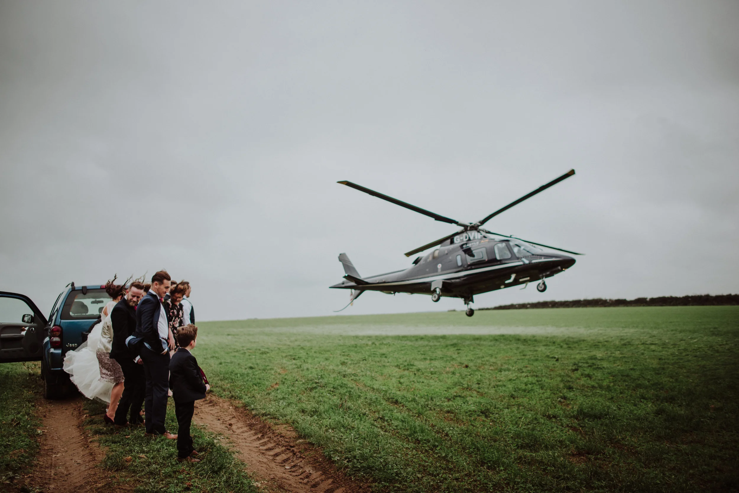 A group of people, including a bride in a wedding dress, waiting on a dirt path near a black helicopter in an open field during overcast weather.