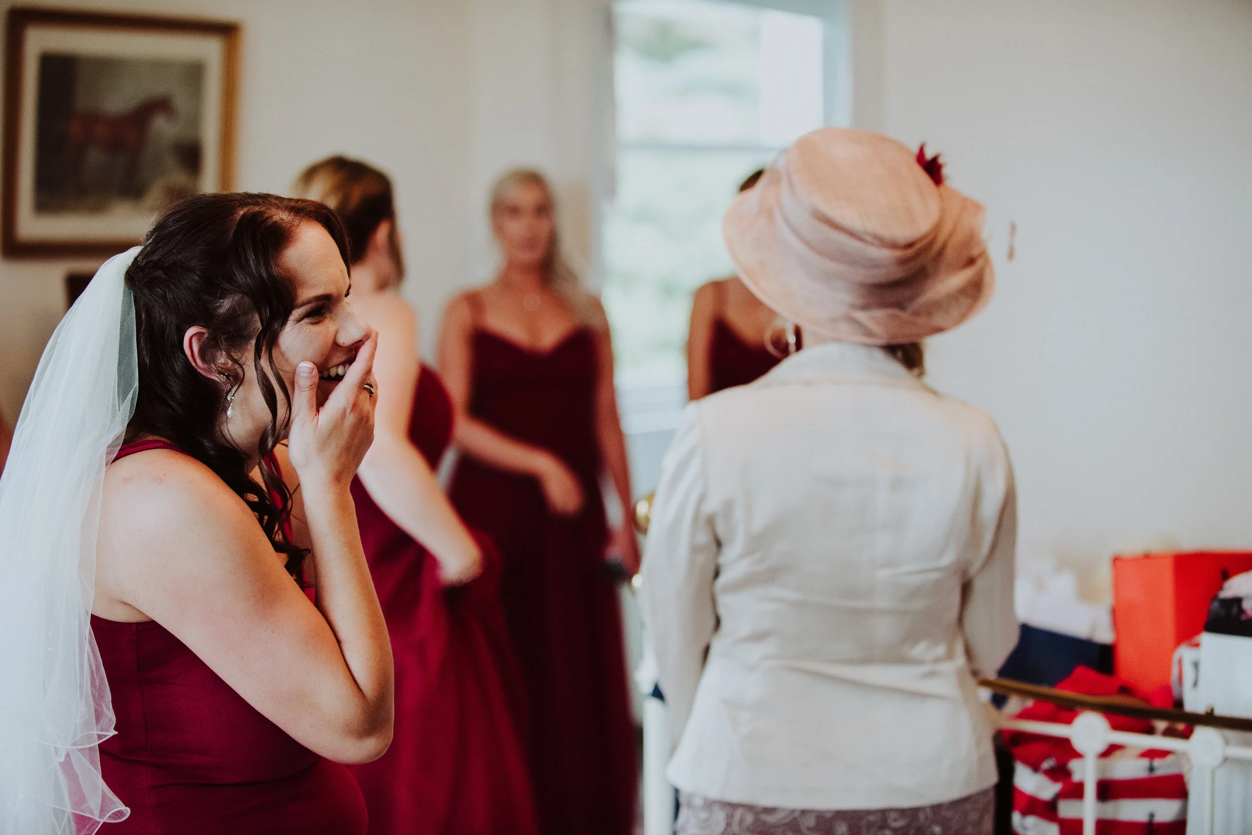 Bride in a white dress and veil laughing with hand covering her mouth, surrounded by bridesmaids in dark red dresses, and a woman in a large beige hat and white jacket, in a room with a painting and a window.