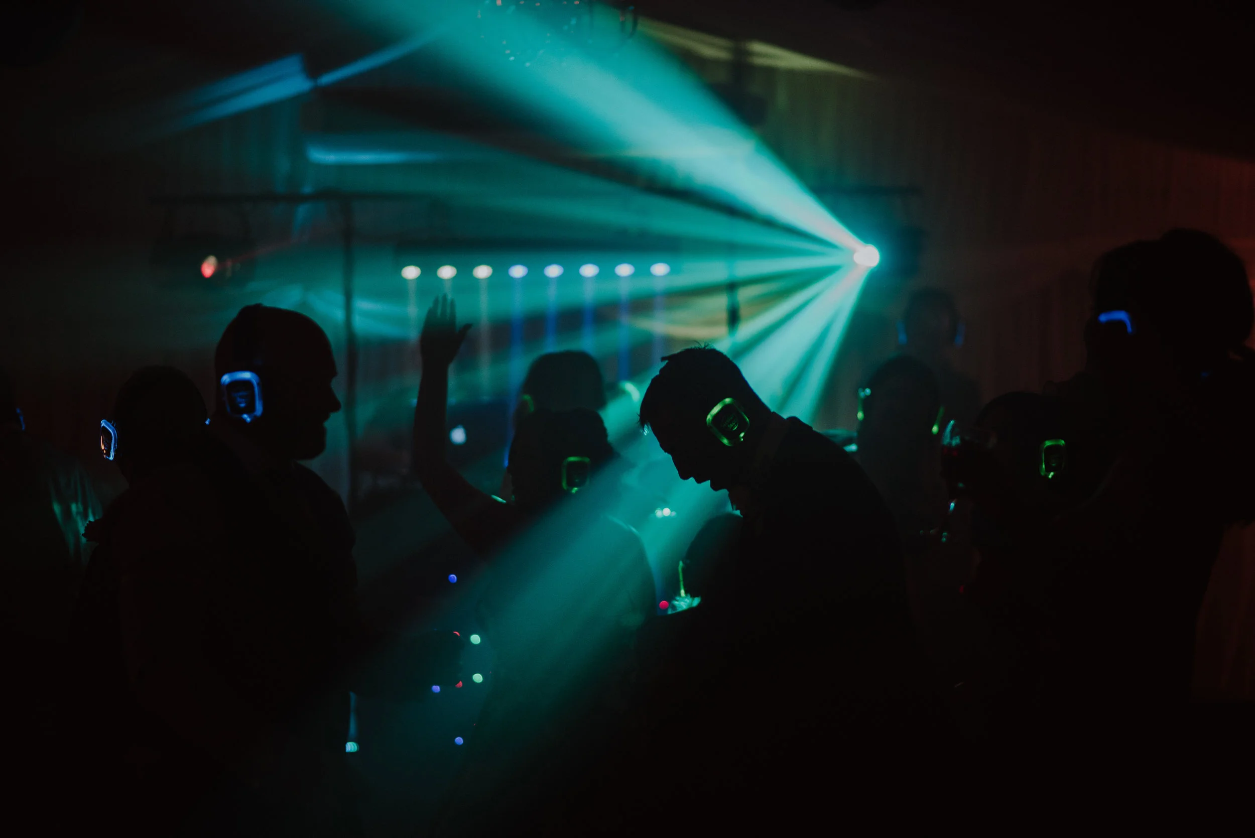 Silhouettes of people wearing headsets at a dance party with colorful laser lights.