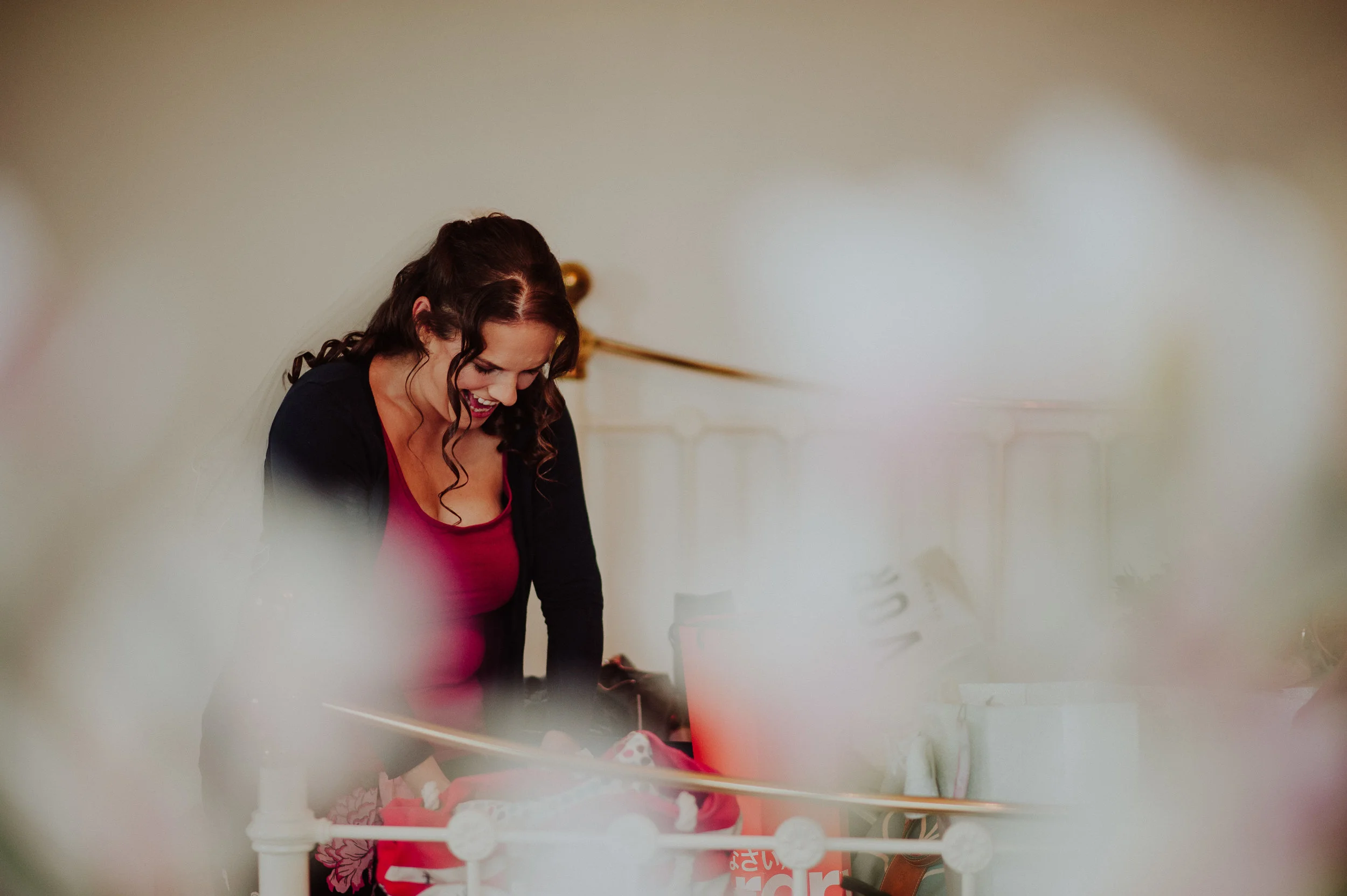 A woman with dark, curly hair, wearing a black jacket and a red top, is smiling and leaning forward, looking down at something, possibly on a bed, in a bedroom with cream-colored walls and a brass bed frame.