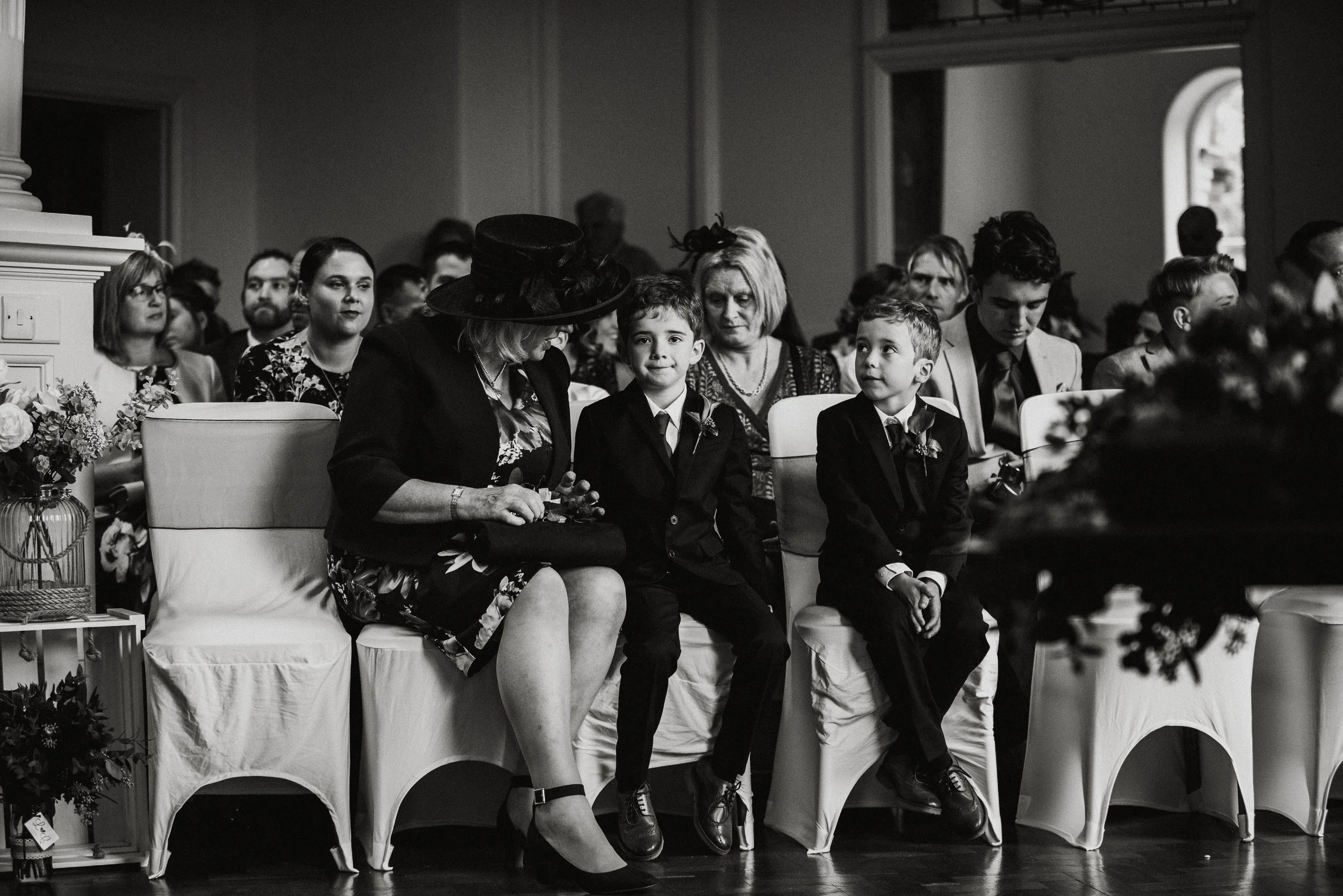 Black and white photo of a diverse group of people sitting in chairs at a formal event, including women, men, and children dressed in suits and dresses, some with flower boutonnieres. They are facing forward, engaged in a ceremony or celebration.