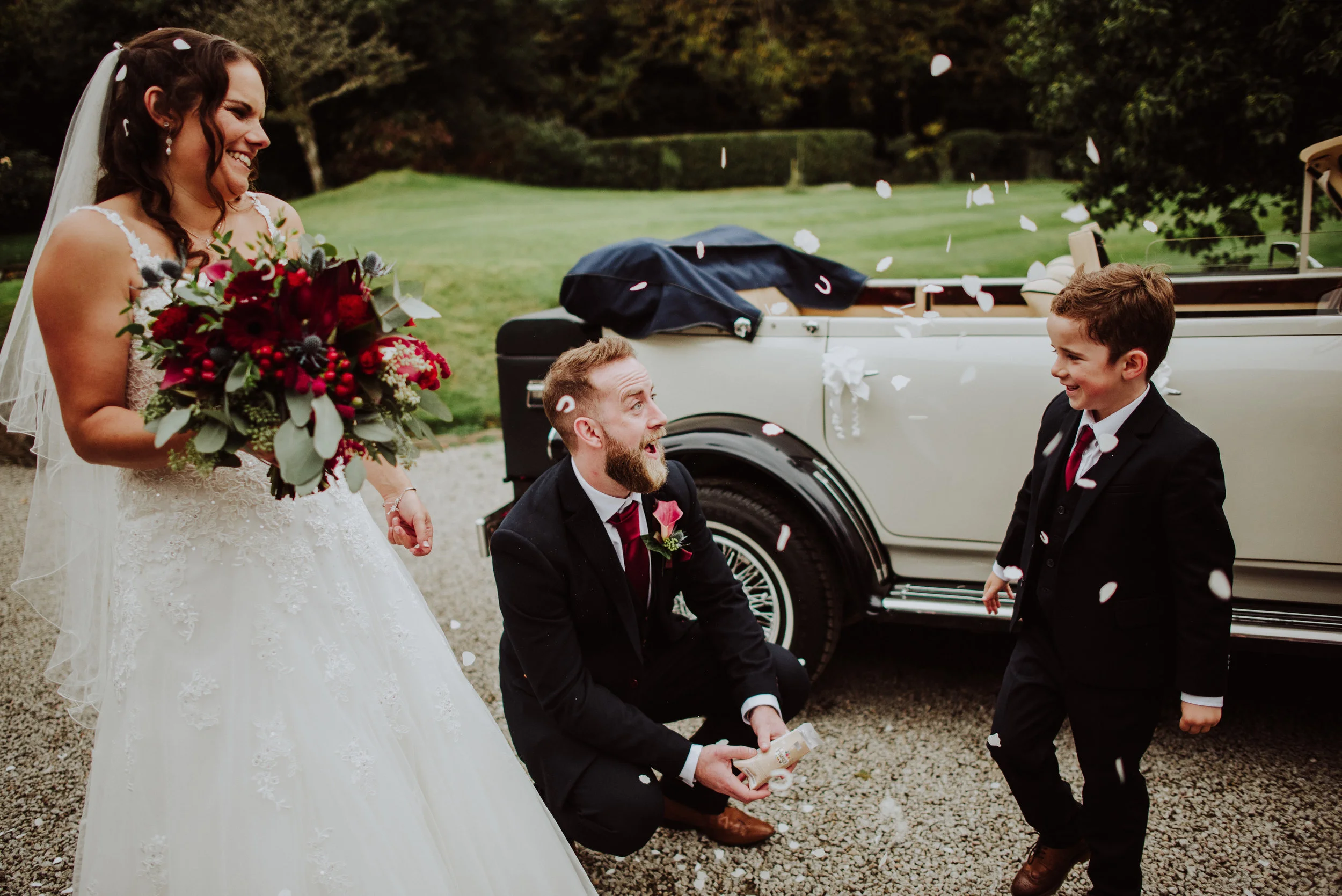 A bride holding a bouquet of red and green flowers, smiling as a man and a young boy in tuxedos celebrate beside her, with white confetti falling around them near a vintage car in a green outdoor setting.