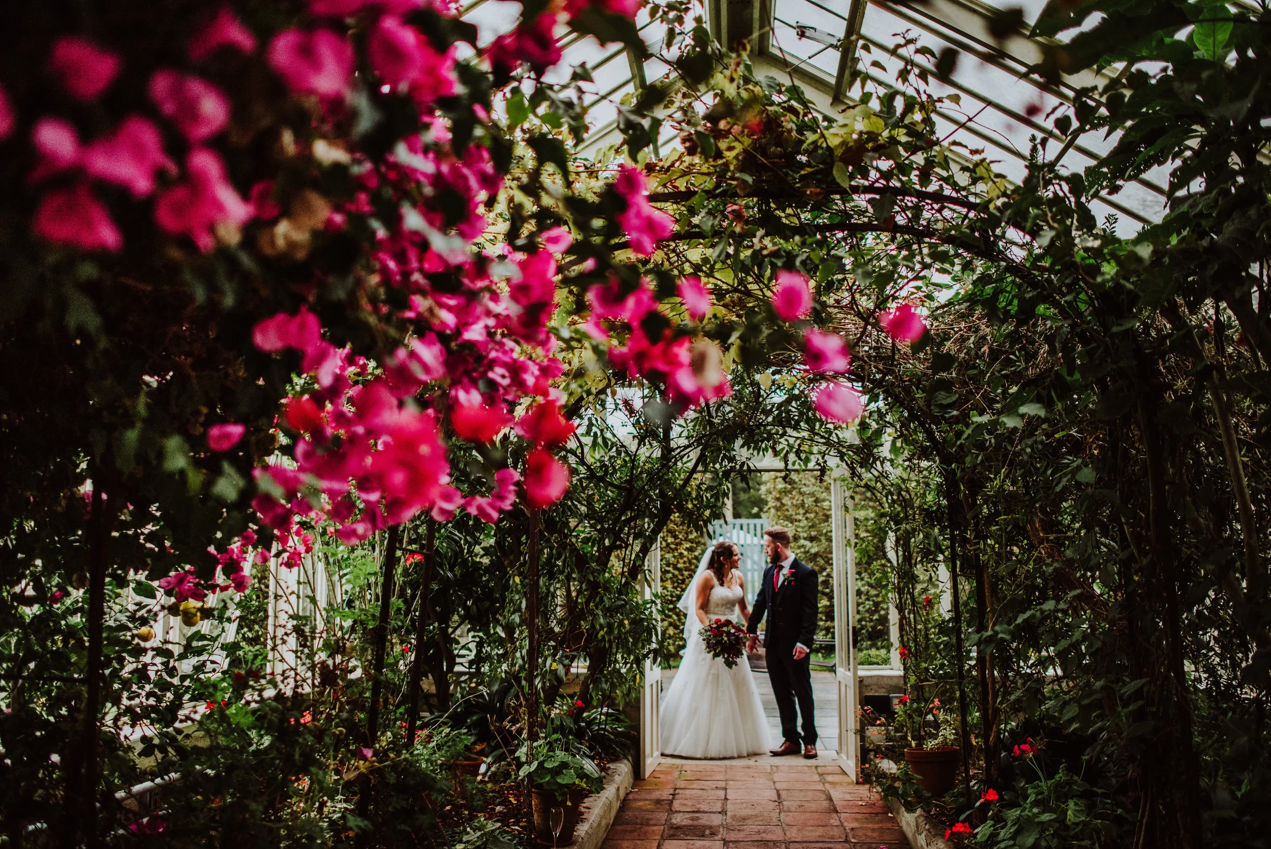 A bride and groom stand together in a garden archway decorated with pink flowers and greenery, holding hands and gazing at each other, with a glass greenhouse structure overhead.