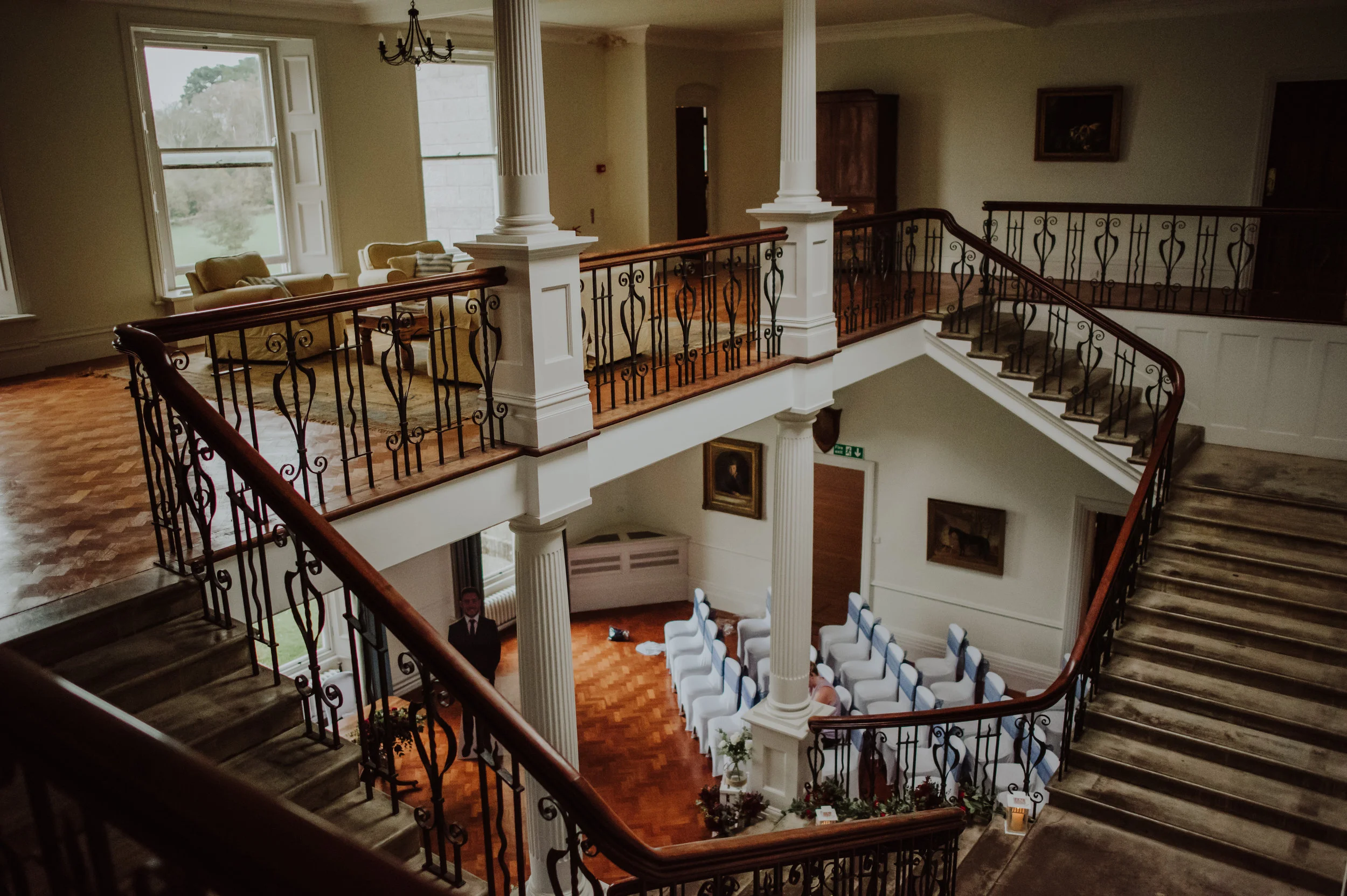 Interior of a historic building with a staircase, balcony, and seating area. The staircase has ornate black iron railings and wooden handrails, leading to an upper floor. The lower level features white chairs arranged for an event, floral arrangement