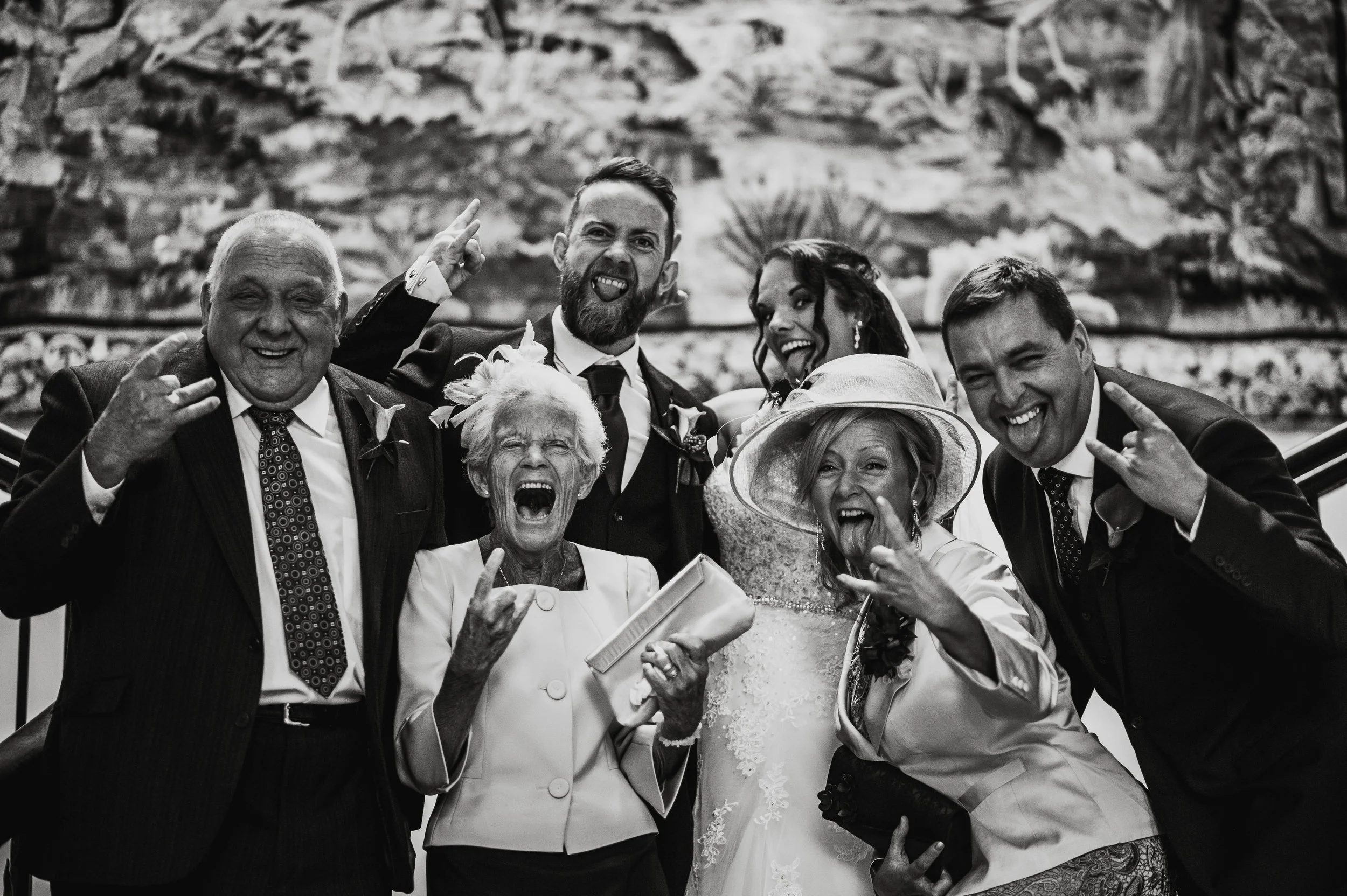 Group of people celebrating at a wedding, including elderly women and men, a bride, and a groom, making cheerful and playful gestures, with a scenic backdrop.