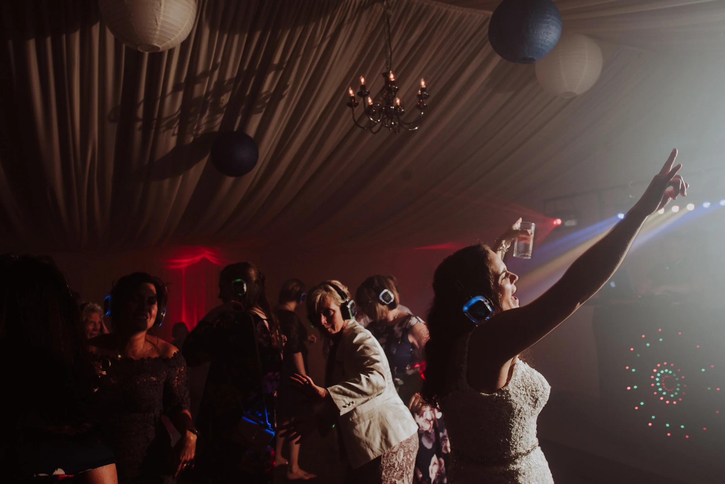 Women dancing at a party with headphones, a woman in the foreground holds a drink and points upward, colorful lights and a chandelier are visible in the background.