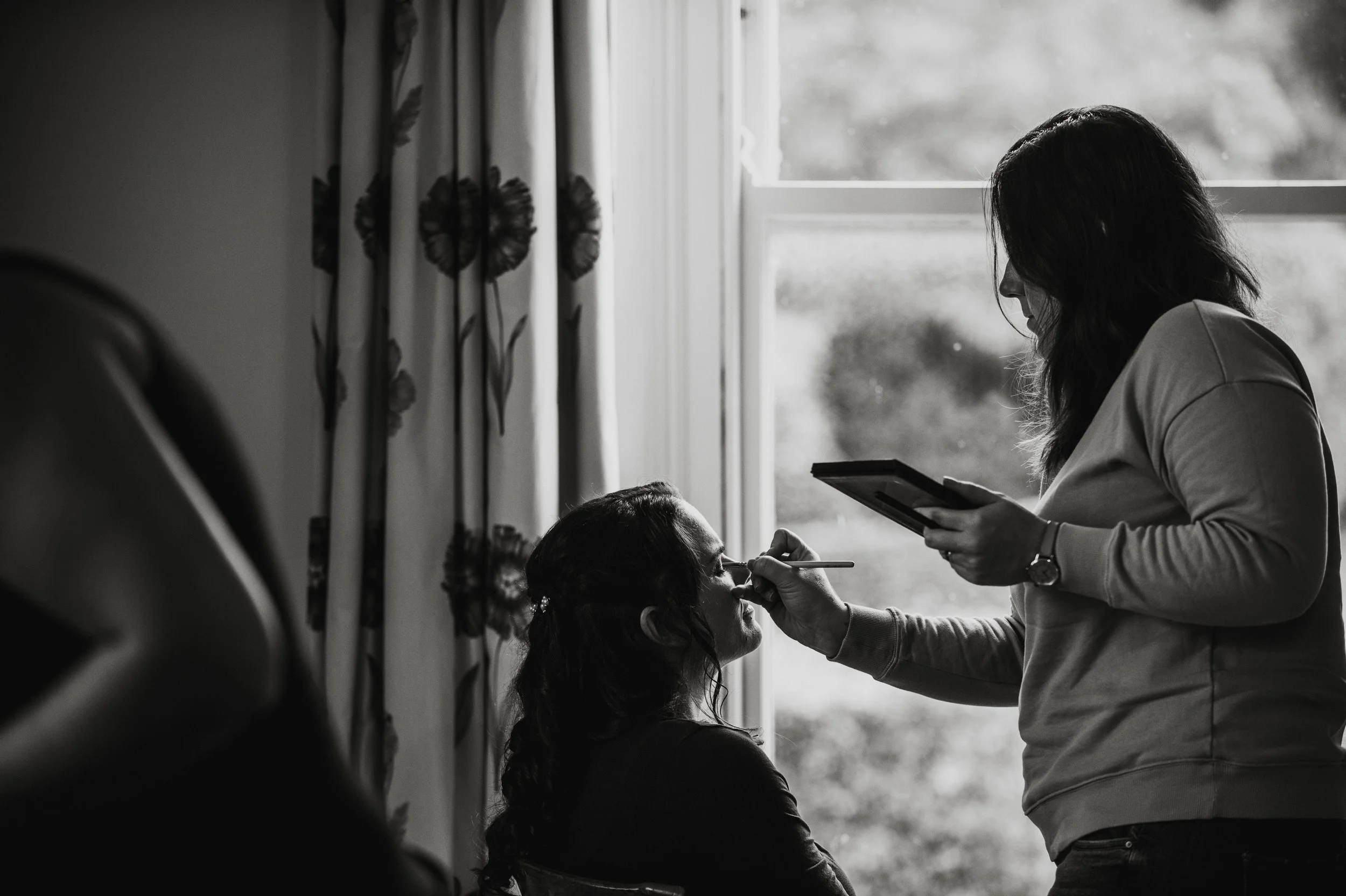A woman applying makeup to another woman seated by a window, while holding a palette in her left hand. The scene is in black and white.