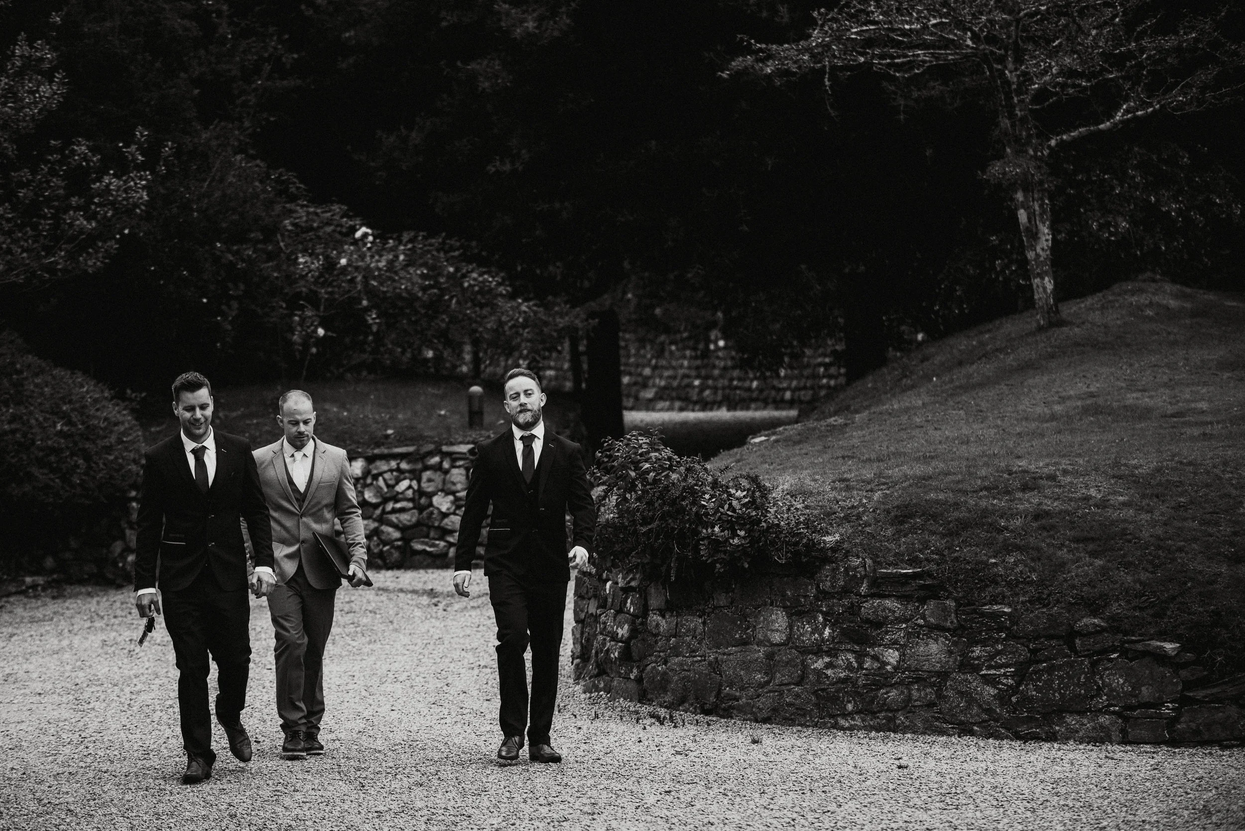 Black and white photo of three men in suits walking outdoors on a gravel path, with trees and a stone wall in the background.