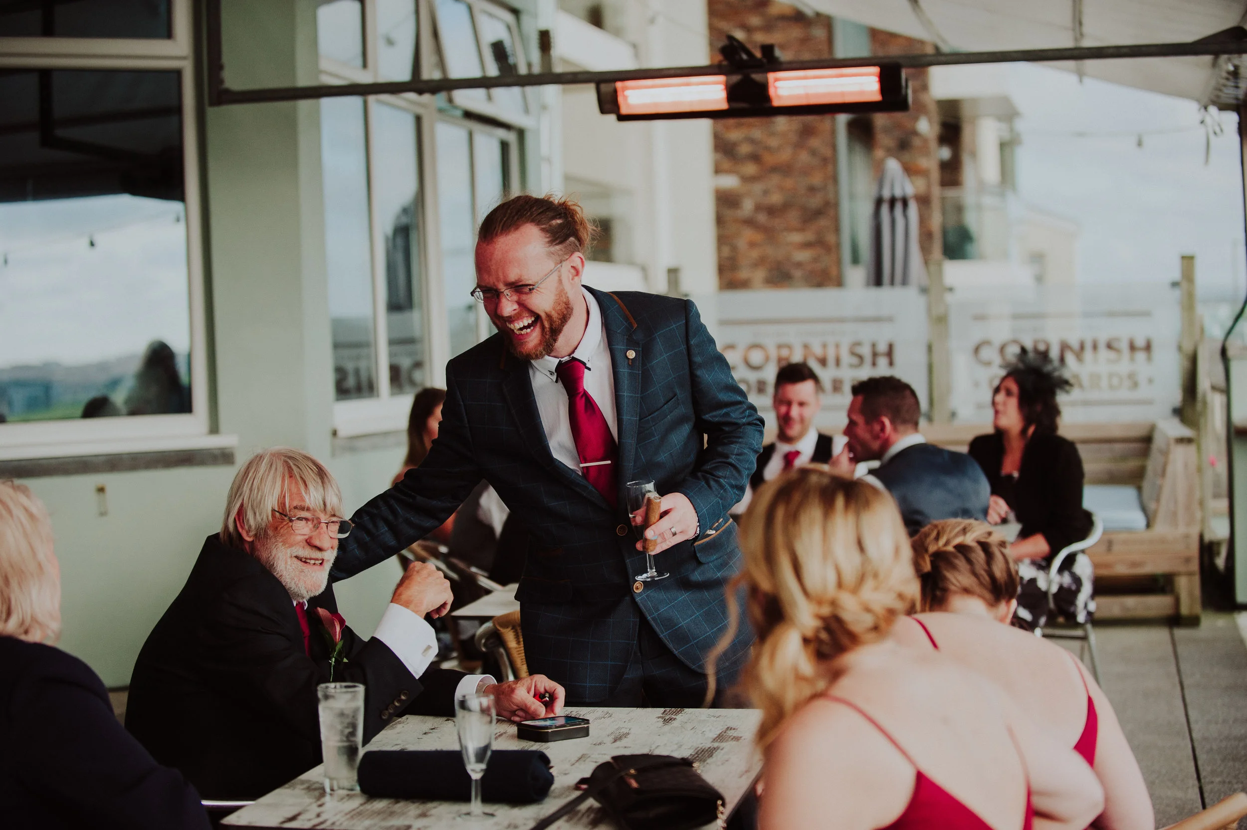 Man in a dark blue checkered suit and red tie smiling and handing a glass to an older man with gray hair and glasses, sitting at a table with guests during a celebration or wedding reception.