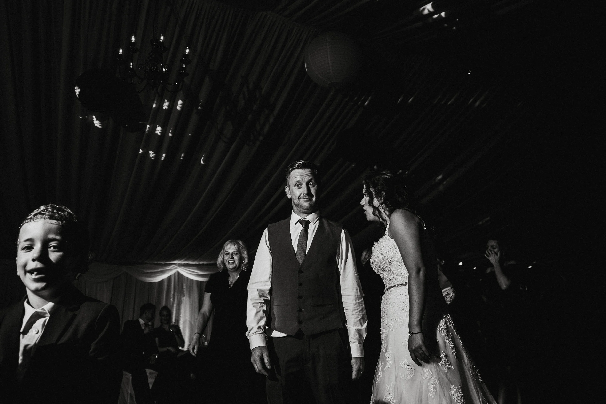 Black and white photo of a wedding reception with several guests. A man in a vest and tie stands in the center, looking at a woman in a wedding dress. A woman with short hair smiles in the background, and a young boy in a suit is in the foreground sm