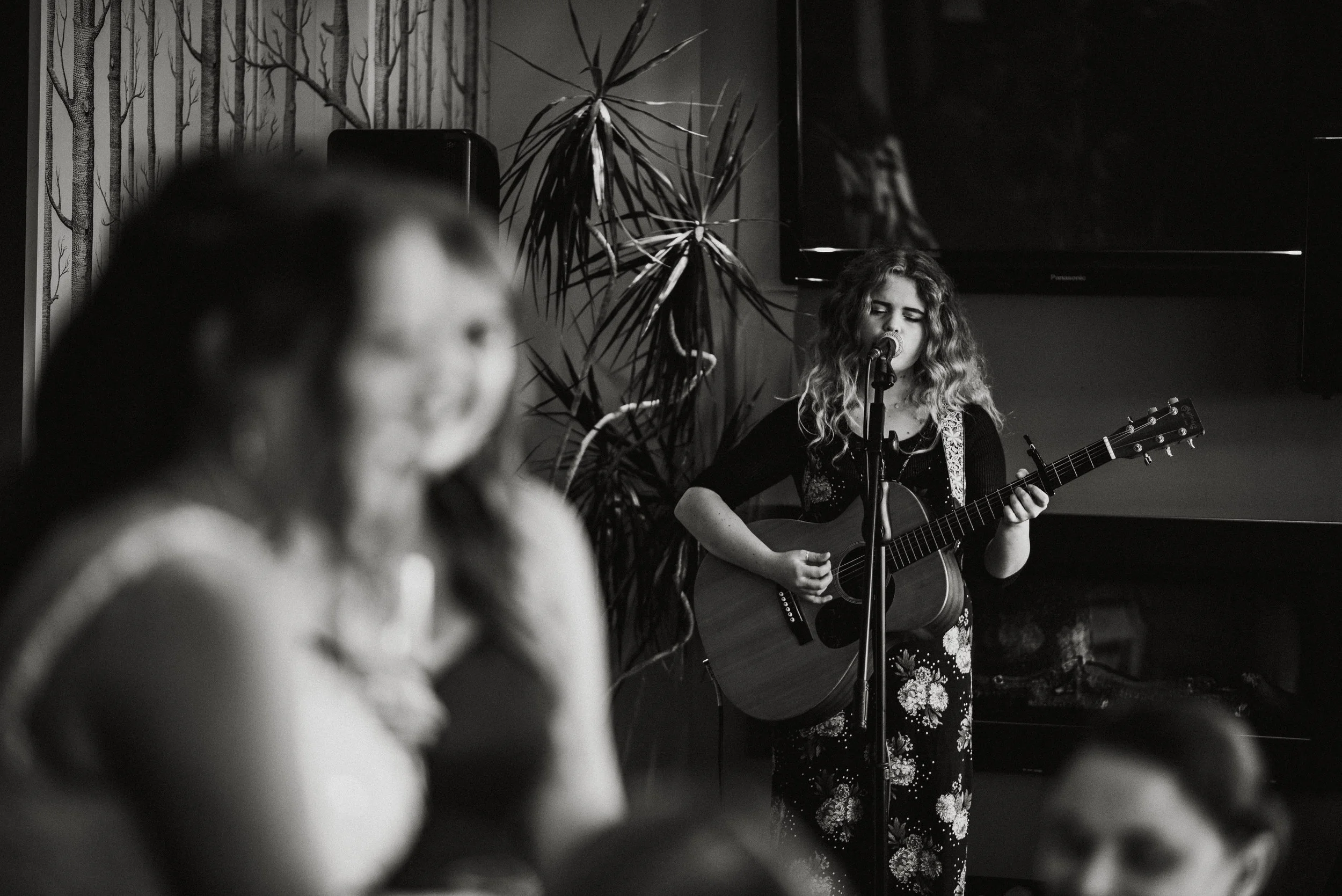 A woman with curly hair singing and playing an acoustic guitar at a microphone on a stage, with dappled lighting, and a woman in the foreground smiling and listening.
