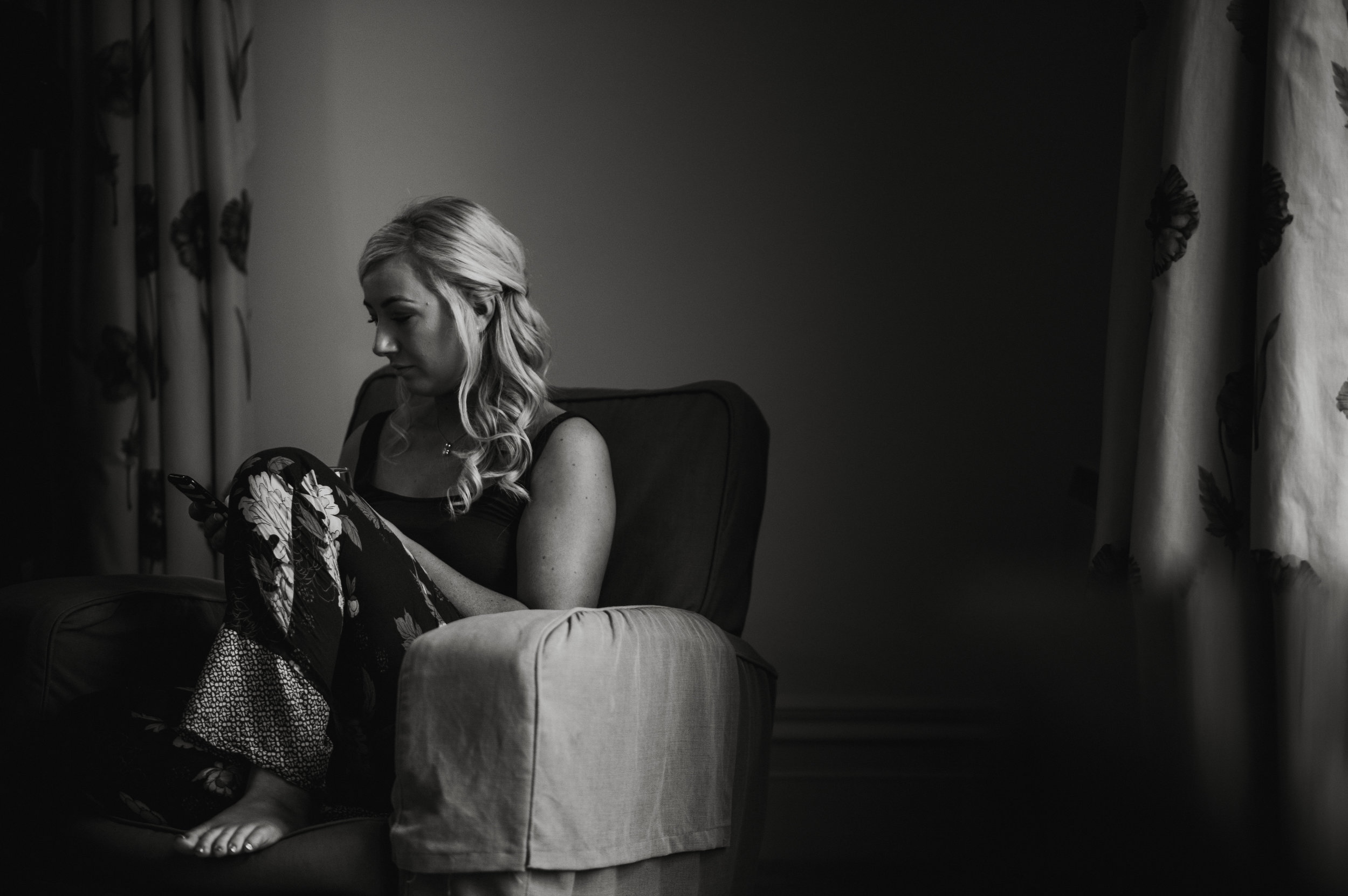 A woman with long, wavy blonde hair, sitting on a cushioned armchair, looking at her phone in a dimly lit room with floral curtains.