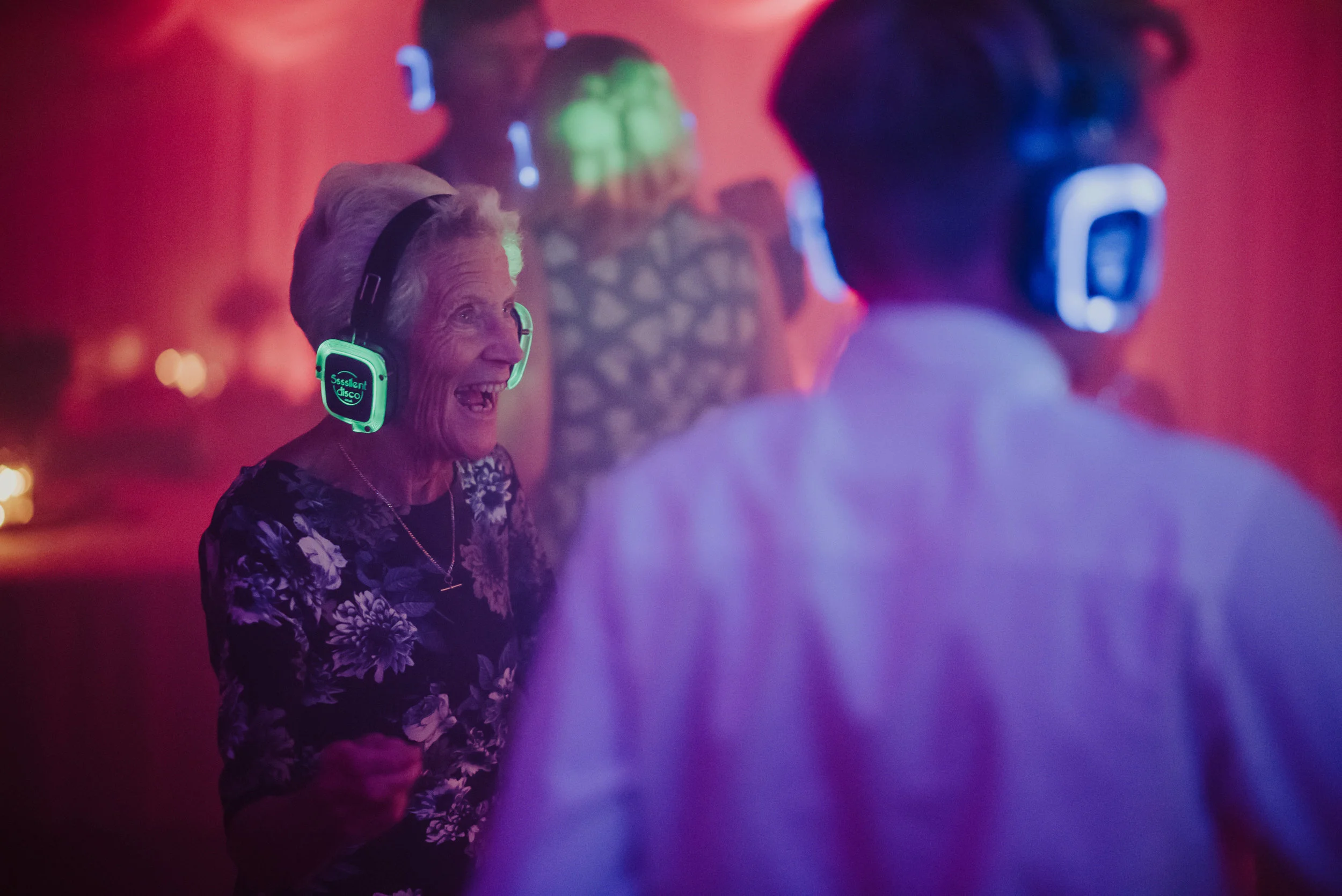 An elderly woman and a man dancing and enjoying music at a party, wearing illuminated headphones in a colorful, lively environment.