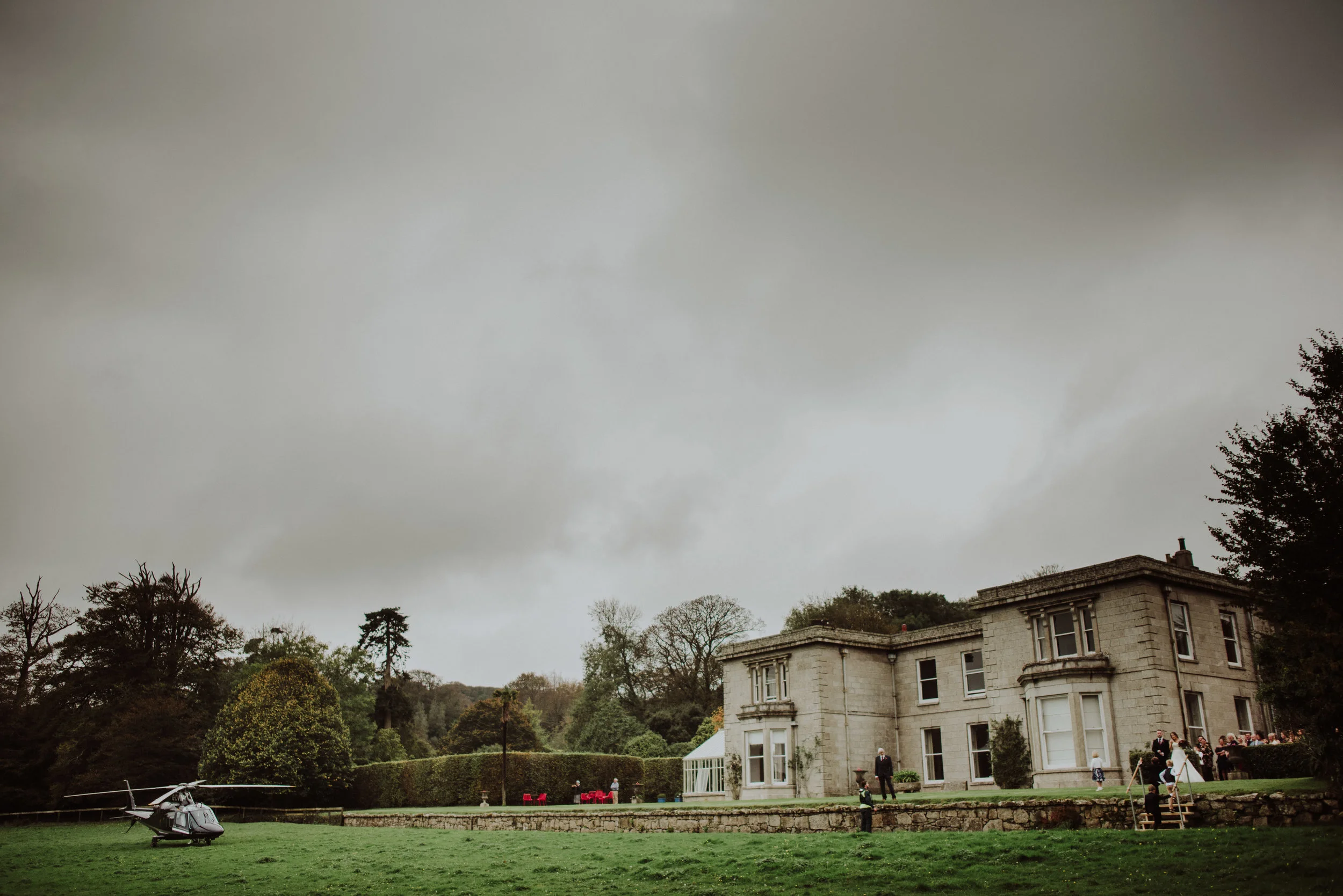 Large estate with a formal garden, a helicopter on the lawn, and a group of people dressed for a wedding near the house in overcast weather.