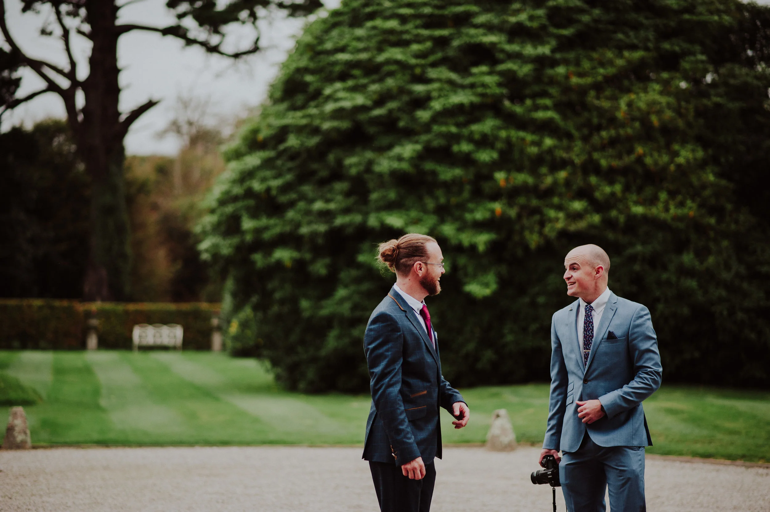 Two men in formal suits conversing outdoors on a gravel path, with lush green trees and grass in the background, one holding a camera.
