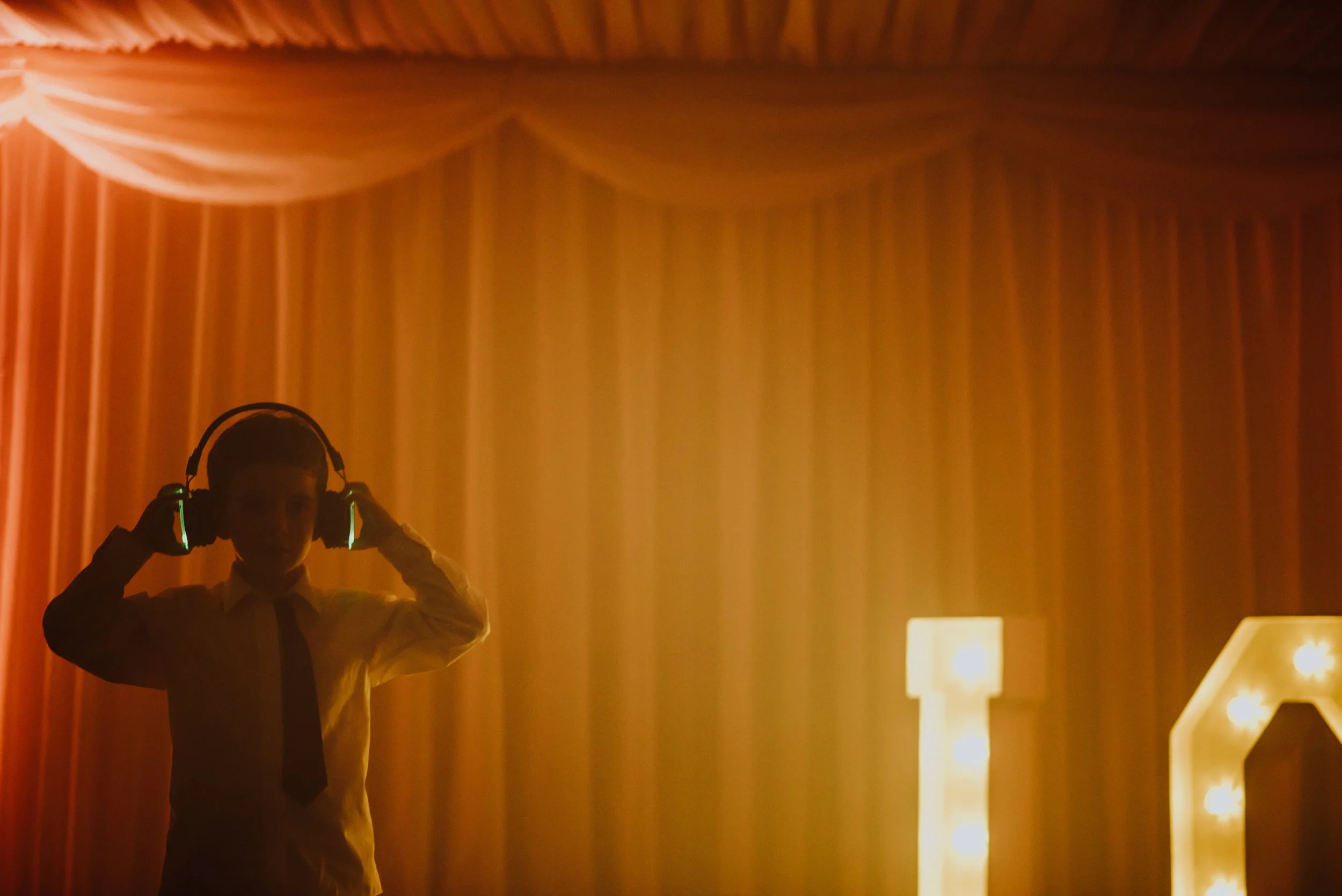 A young boy in formal attire, wearing a dress shirt and tie, is adjusting his glowing headphones in front of a curtain with warm lighting; illuminated letters are partially visible on the right side.