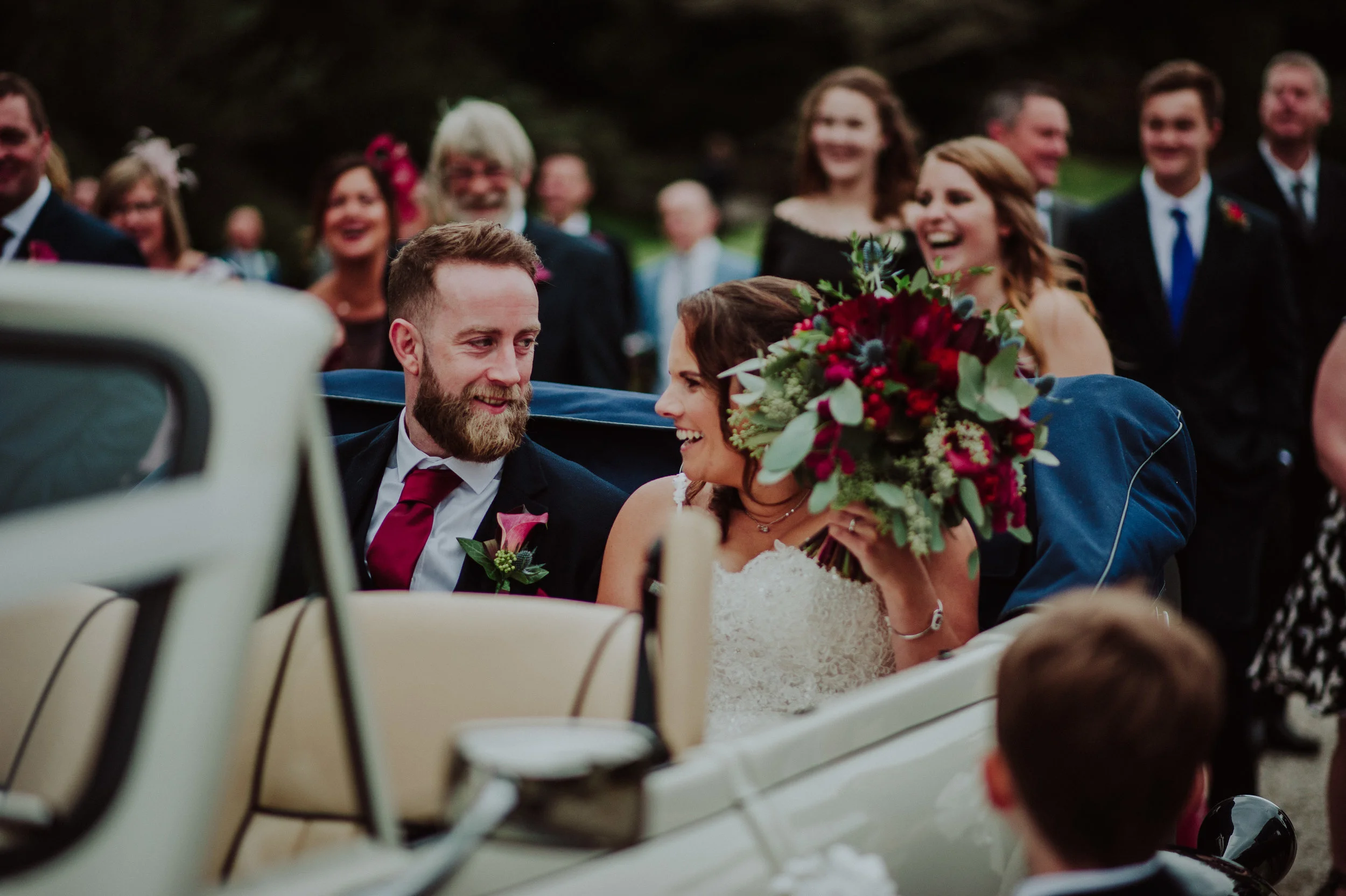 Bride and groom sitting in a vintage car surrounded by wedding guests, smiling and laughing, bride holding a large bouquet of red and green flowers.