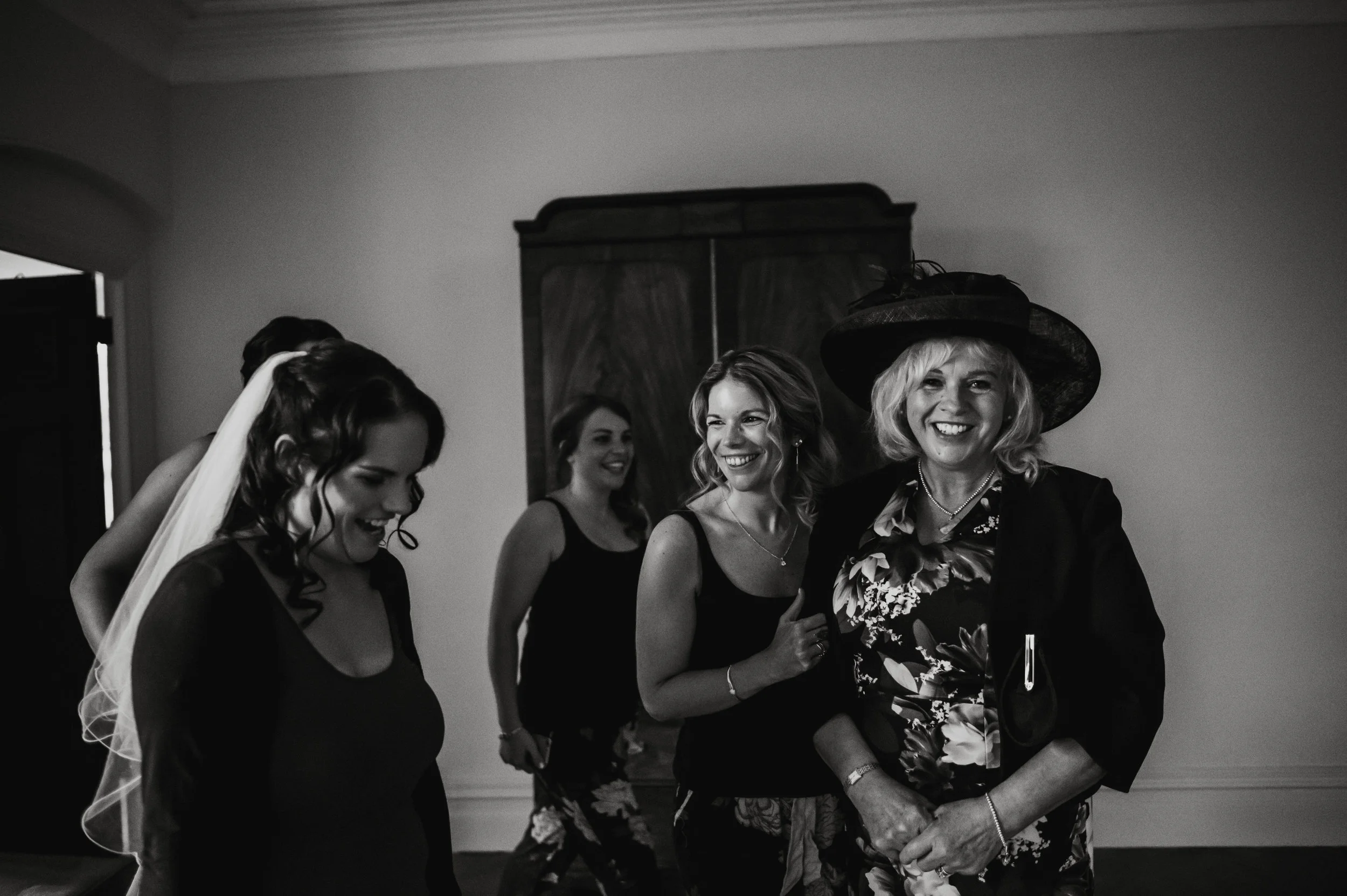 Four women smiling and talking in a room, with one woman wearing a large hat and a floral shirt, and a mirror in the background.