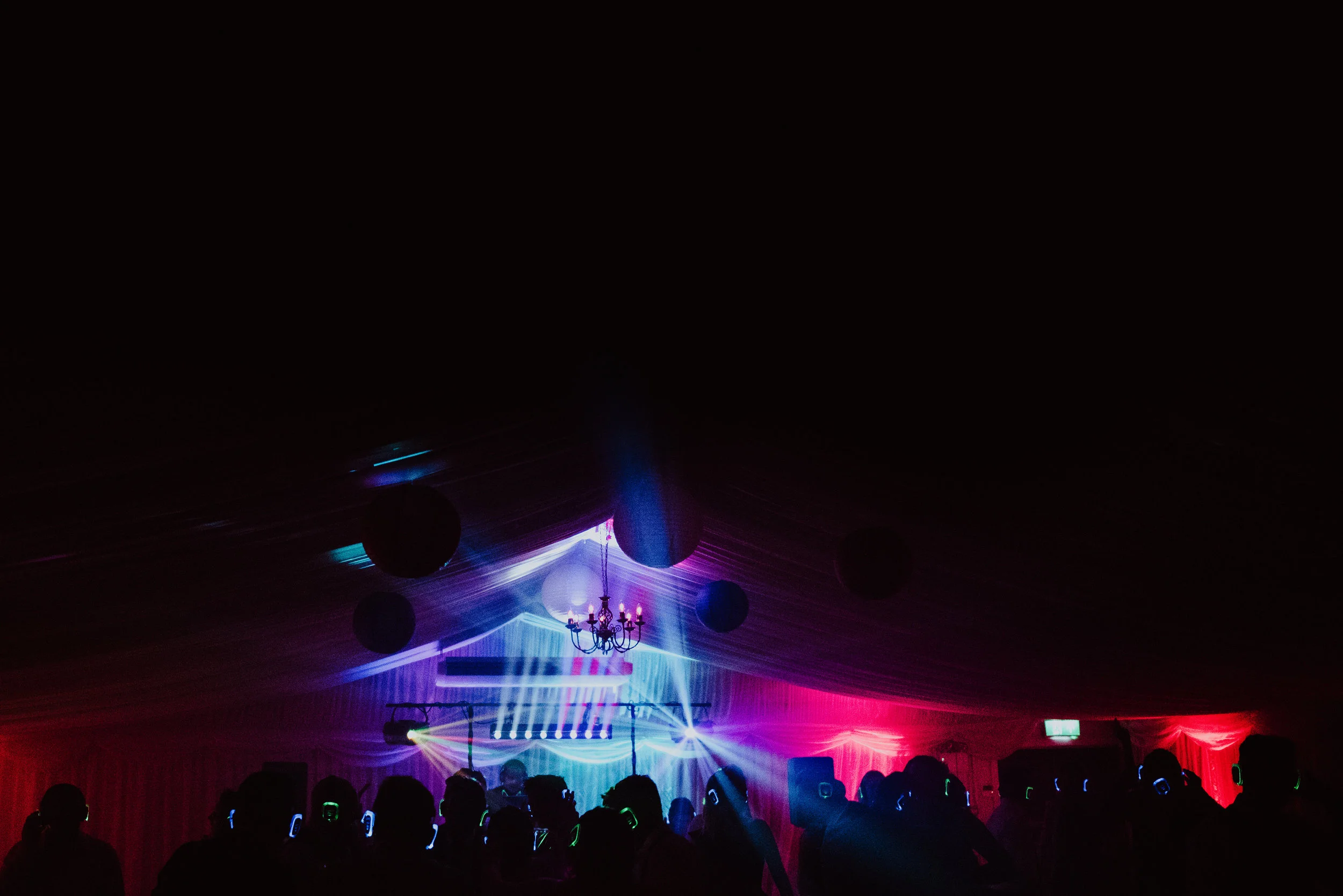 Party scene with silhouettes of people wearing headphones, colorful lights and laser effects, chandeliers, and a DJ booth.