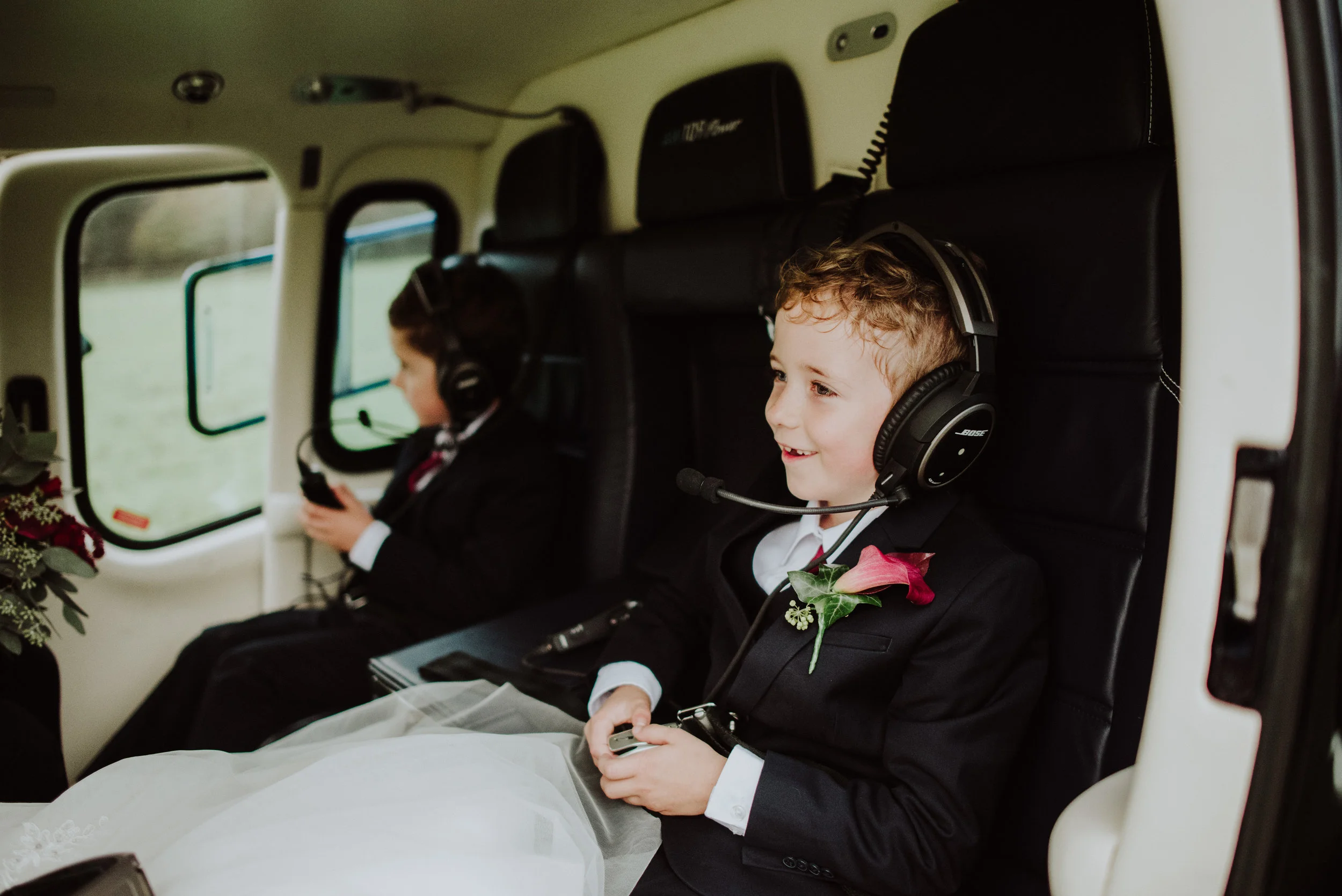 Two children dressed in formal wedding attire sitting inside a helicopter. The boy in the foreground, smiling and holding a device, has a pink flower boutonniere. The girl in the background is looking at her phone, both wearing headsets.