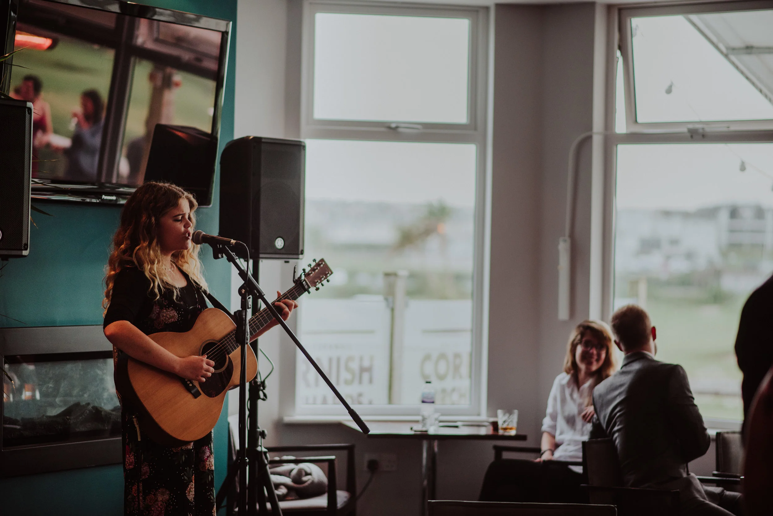 A young woman with curly hair playing an acoustic guitar and singing into a microphone in a room with large windows, with a few people sitting and chatting in the background.