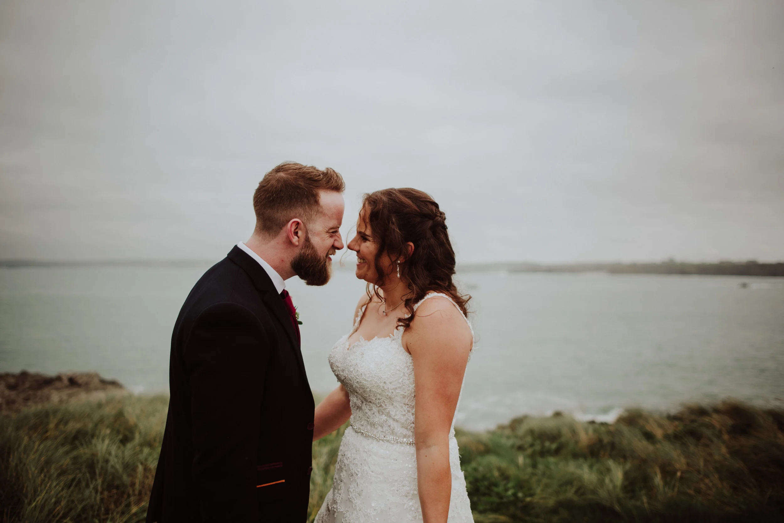 A newlywed couple with foreheads touching, smiling at each other on a beach with water and cloudy sky in the background.