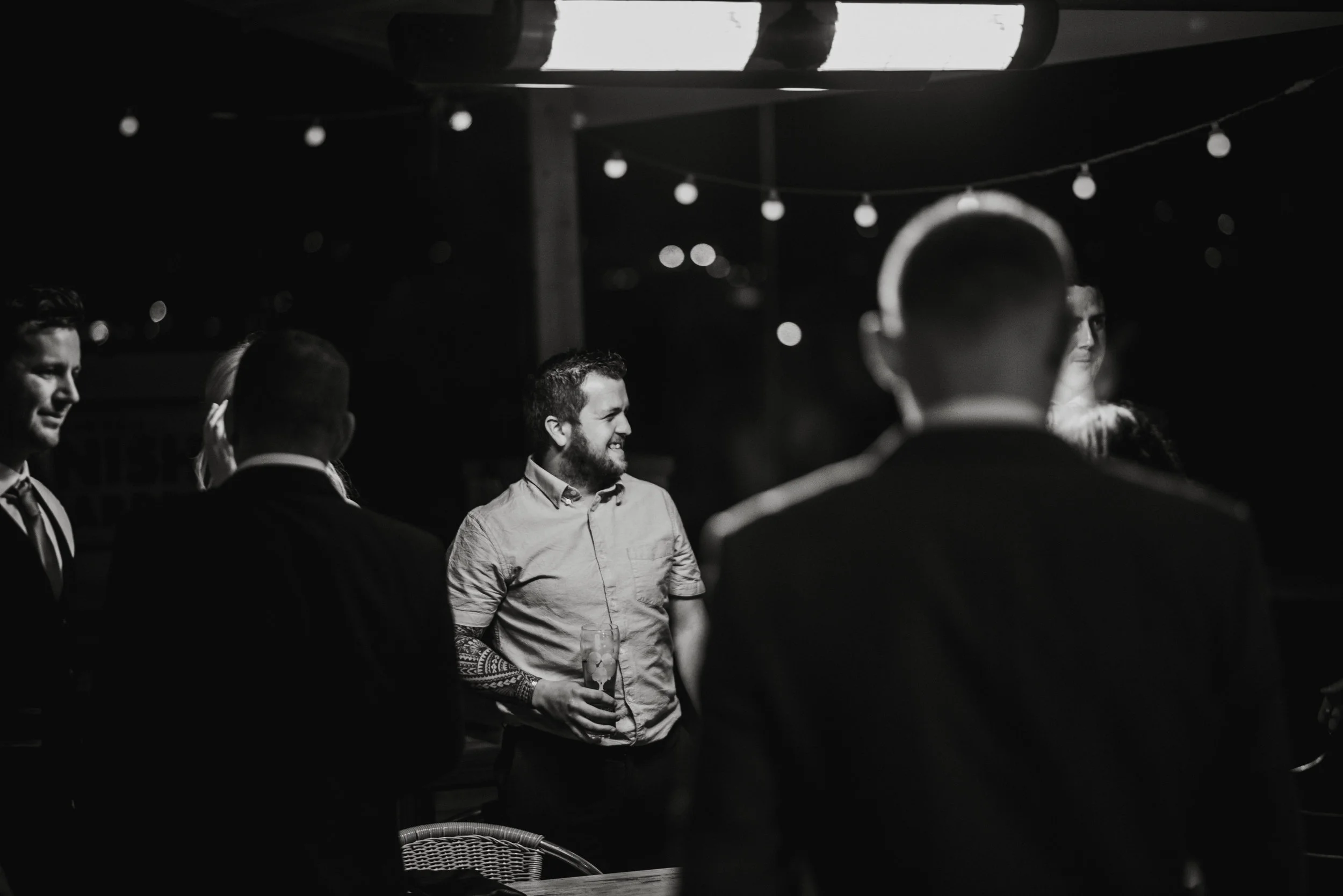 Group of people at a nighttime gathering, with string lights overhead, one man smiling and holding a drink, others in dark suits, in a casual setting.