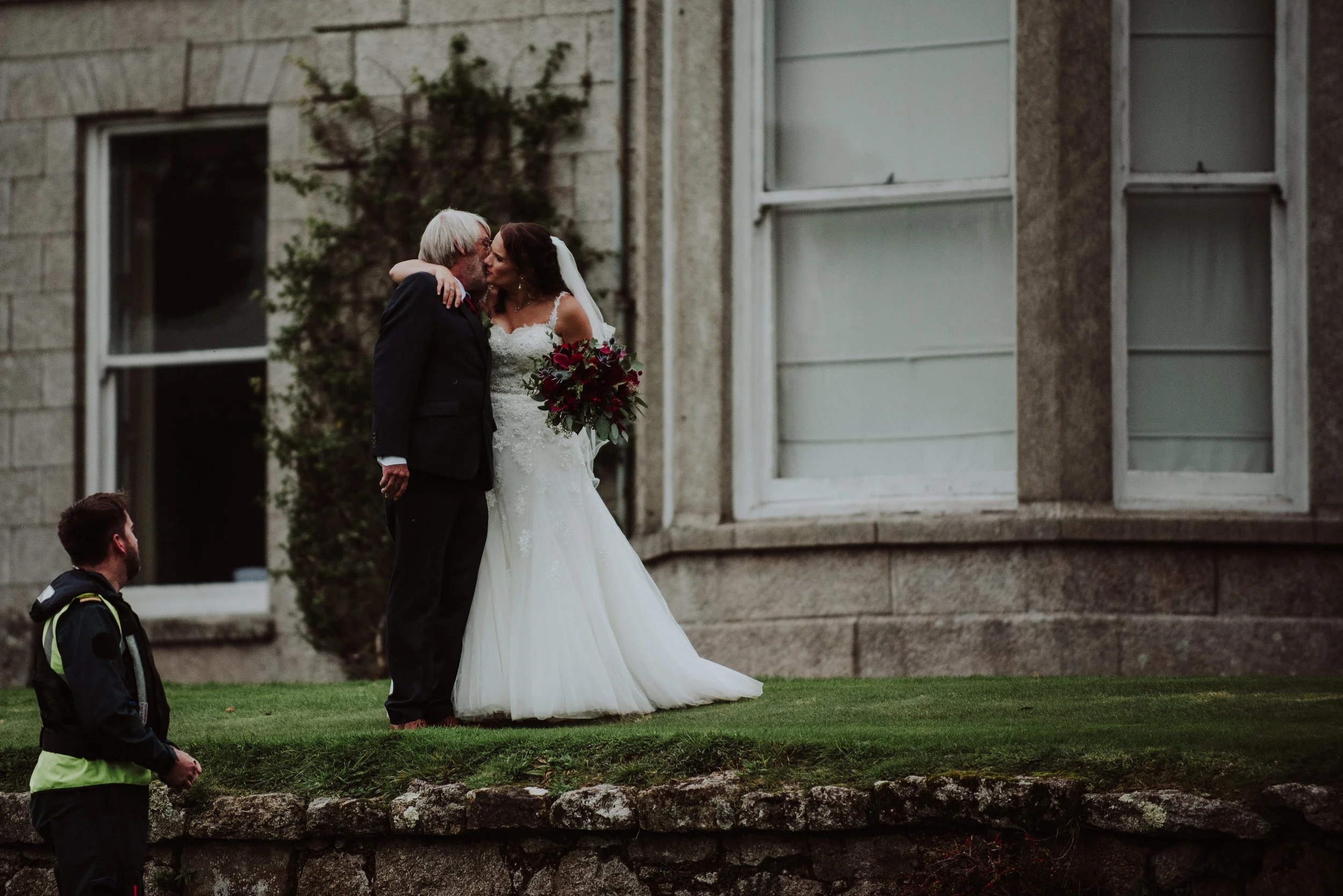 A bride and an elderly man sharing a kiss outside in front of a stone building. The bride is dressed in a white wedding gown and holding a bouquet of flowers. There is a man in a black jacket and yellow vest standing nearby.