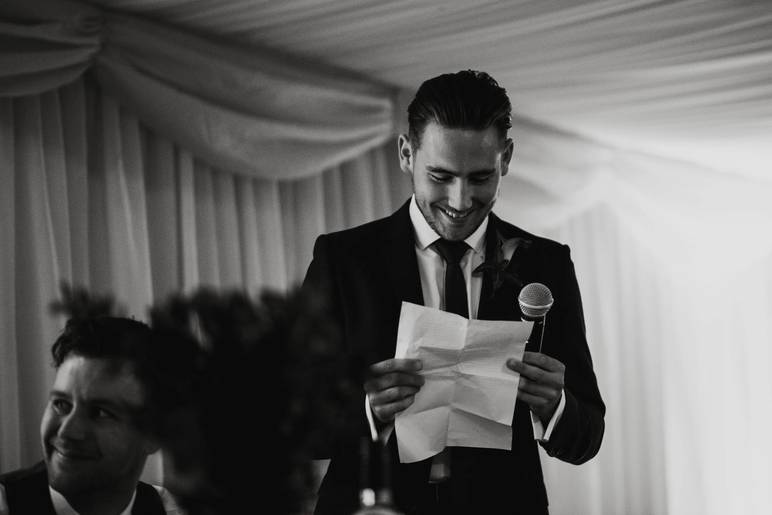 A man in a suit reading a note into a microphone at a formal event, with another man smiling in the background.