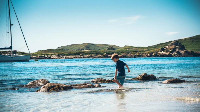 A young boy playing in shallow water near rocks with a sailboat docked in the distance on a sunny day - Isles of Scilly Professional Photographer - Mark Shaw Photography 