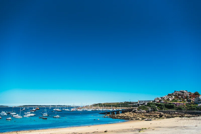 Isles of scilly photography. Beach with boats in the water, rocks and hillside with houses, under a clear blue sky.