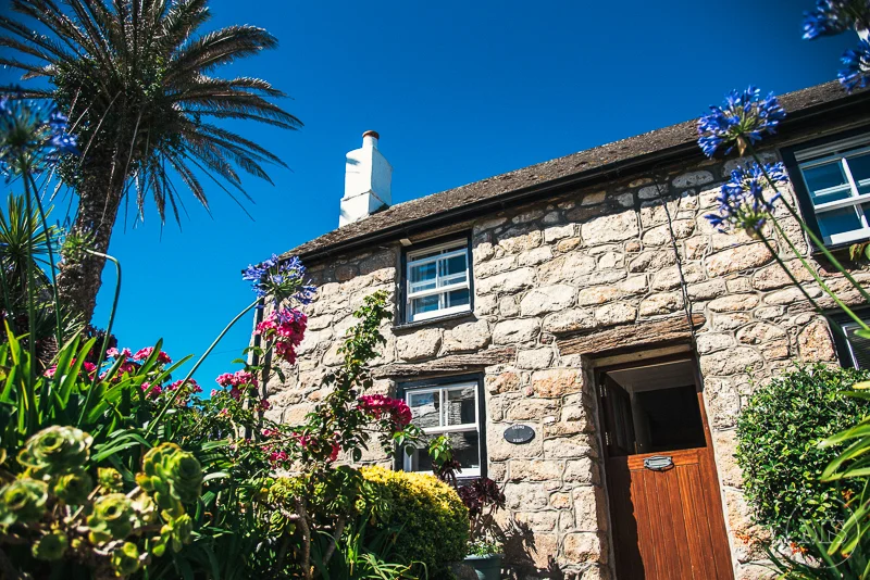 Isles of scilly photography. Stone house with small windows and a wooden door, surrounded by colorful flowers and greenery, with a tall palm tree in the background under a clear blue sky.
