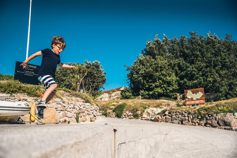 A child jumping off a boat dock onto the pavement on a sunny day with trees and a blue sky in the background.