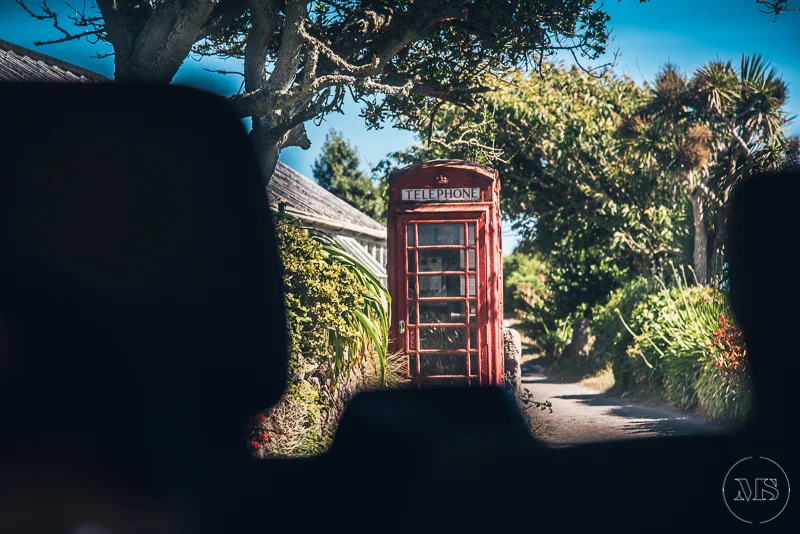 A red vintage telephone booth on a narrow dirt road surrounded by lush greenery and trees, with a clear blue sky overhead.